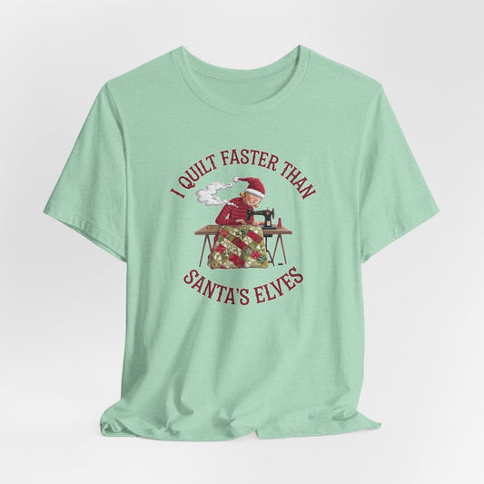 A Heather Mint christmas quilting t-shirt featuring  a quilter working at a sewing machine, quilting a festive holiday quilt, with the phrase "I Quilt Faster Than Santa's Elves" above the image, highlighting the speed and skill of the quilter.