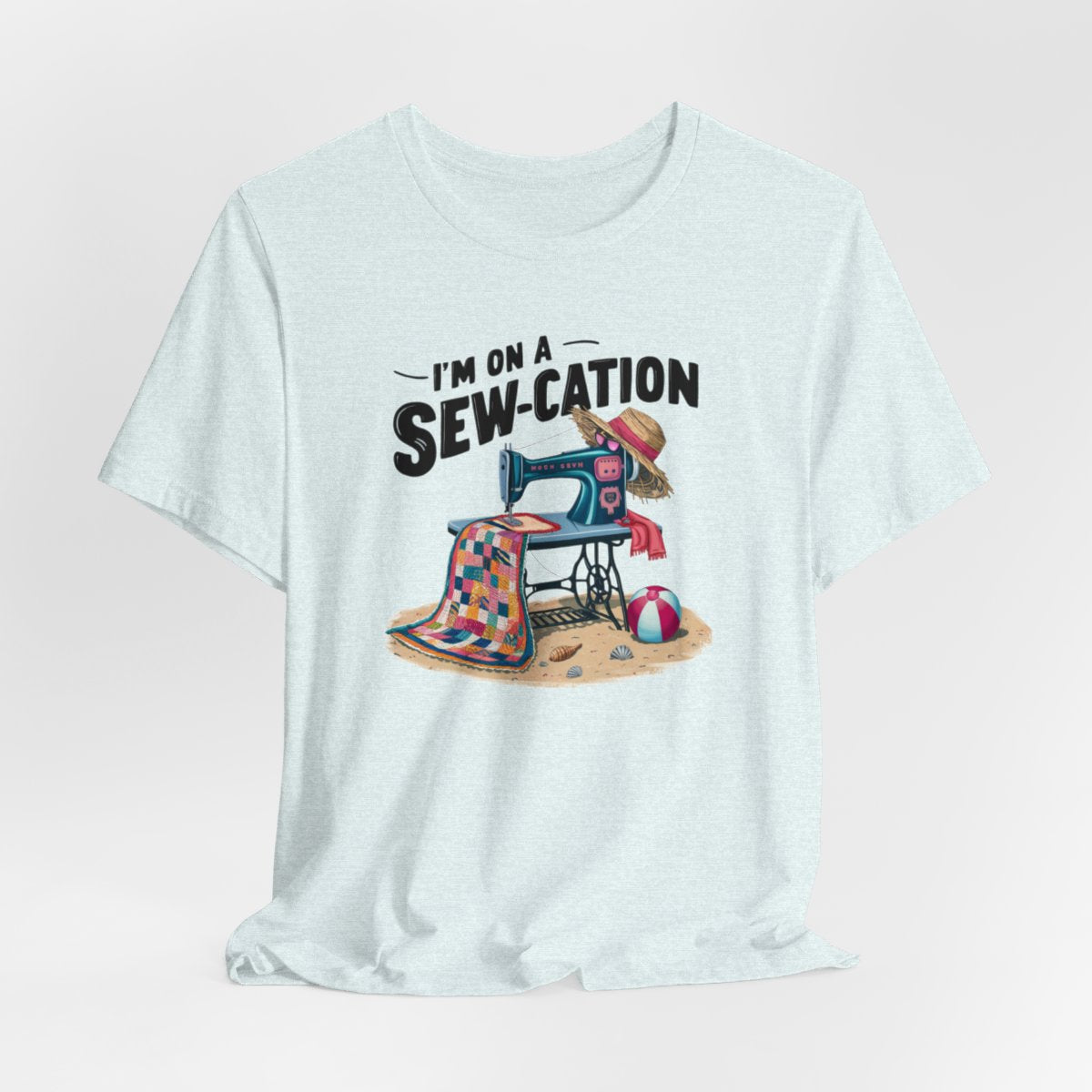 A Heather Ice Blue fun Quilting T-shirt of a vintage sewing machine on a beach with a colorful quilt, sun hat, and beach ball. Text above reads "I'm on a Sew-cation" in bold letters.