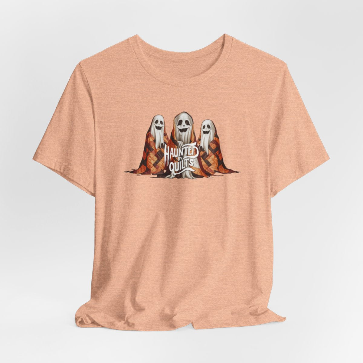 A Heather Peach T-Shirt of three smiling cartoon ghosts covered in colorful patchwork quilts. The quilts feature orange, brown, and black patterns. Text below reads "HAUNTED BY QUILTS" in a spooky white font.