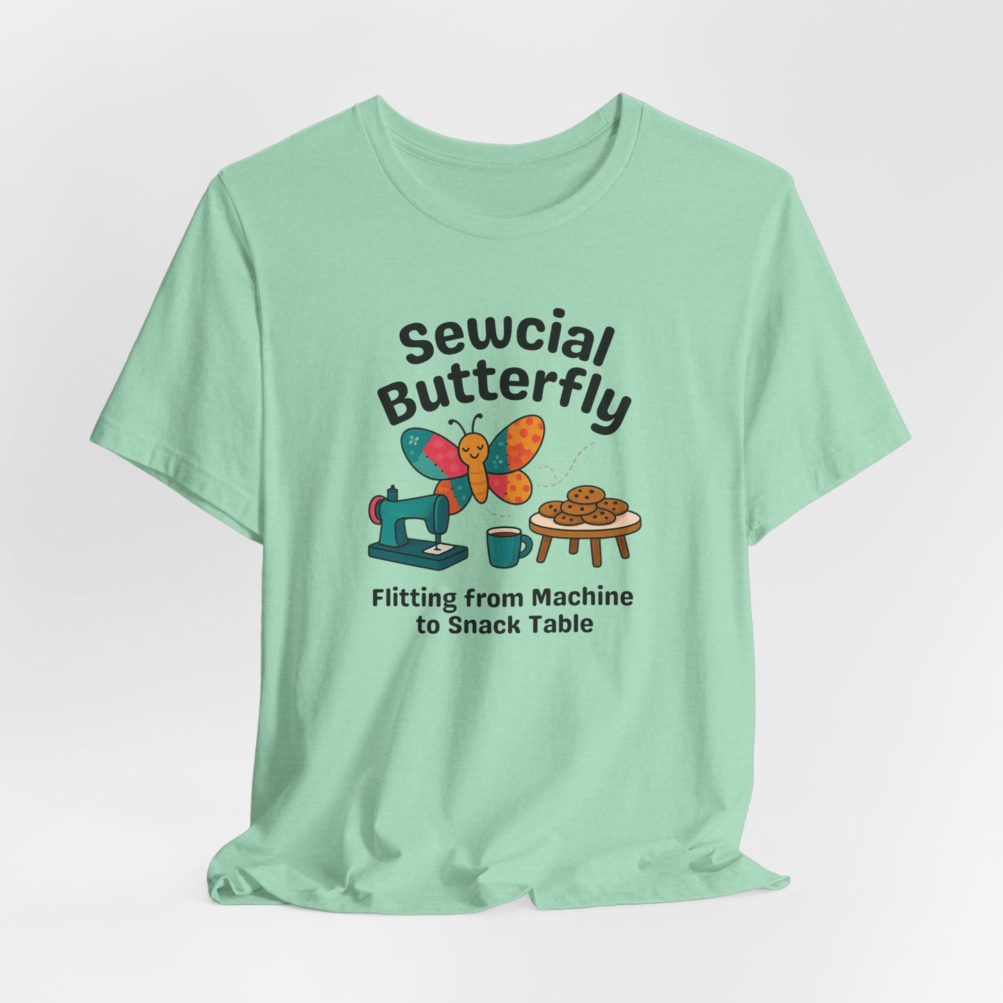 A A Heather Mint t-shirt with a colorful design showing a patchwork butterfly flying between a sewing machine and a table of cookies. Text reads “Sewcial Butterfly – Flitting from Machine to Snack Table.” Funny gift for quilters