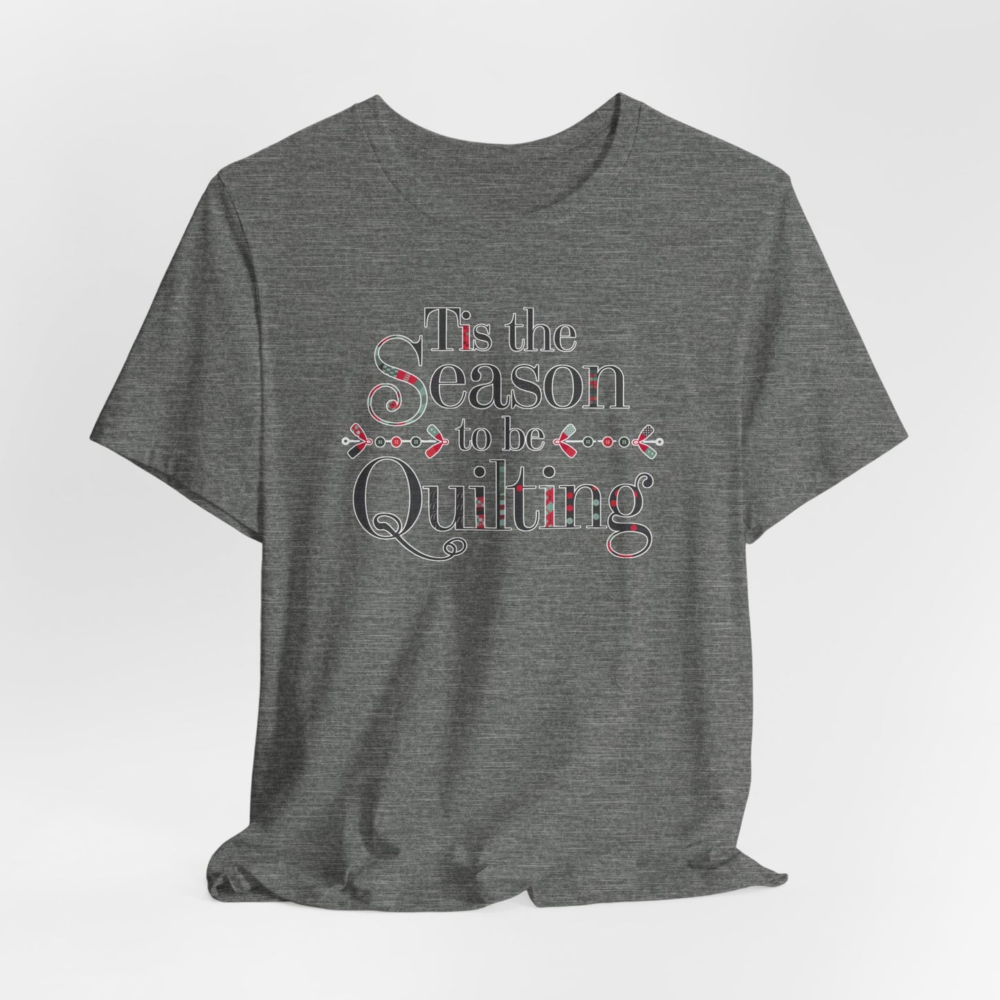 A Deep Heather festive t-shirt featuring the phrase "'Tis the Season to be Quilting," with playful sewing-inspired elements like needles, buttons, and fabric patterns integrated into the typography.