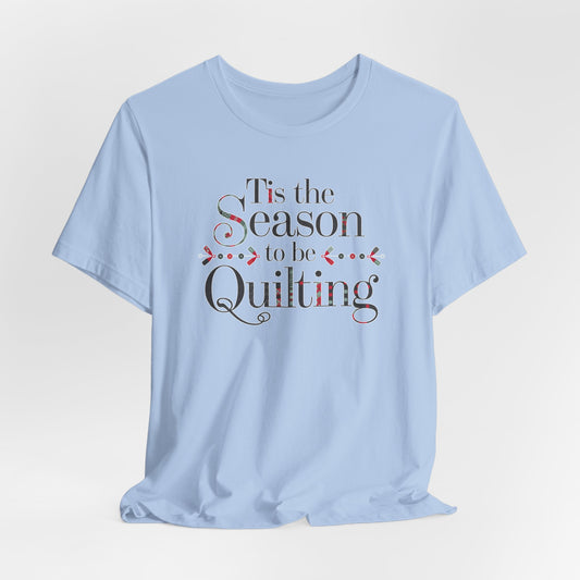A Baby Blue festive t-shirt featuring the phrase "'Tis the Season to be Quilting," with playful sewing-inspired elements like needles, buttons, and fabric patterns integrated into the typography.