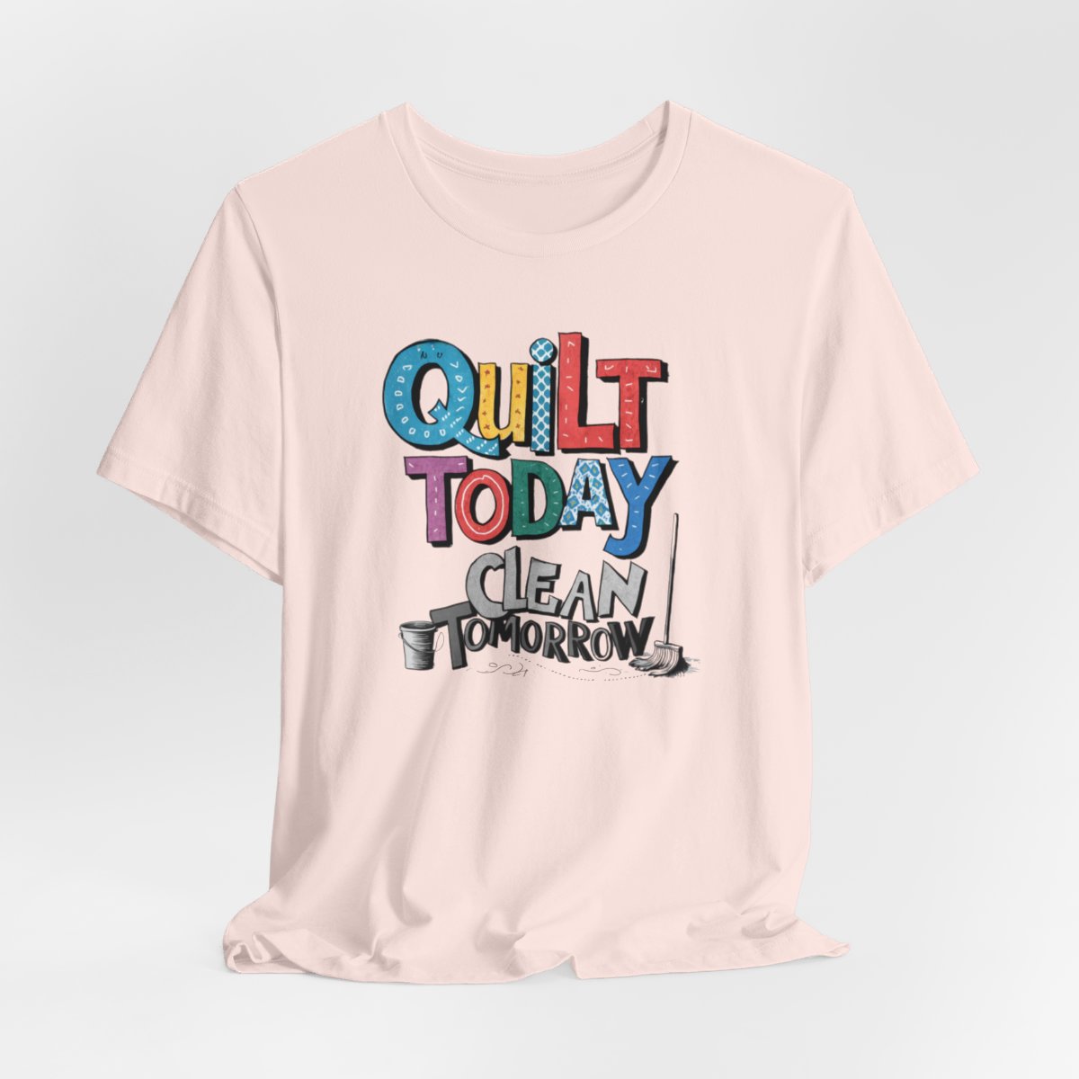 A Soft Pink funny quilting T-Shirt with the phrase Quilt Today, Clean Tomorrow in a bold and cheerful font with a broom off to the side