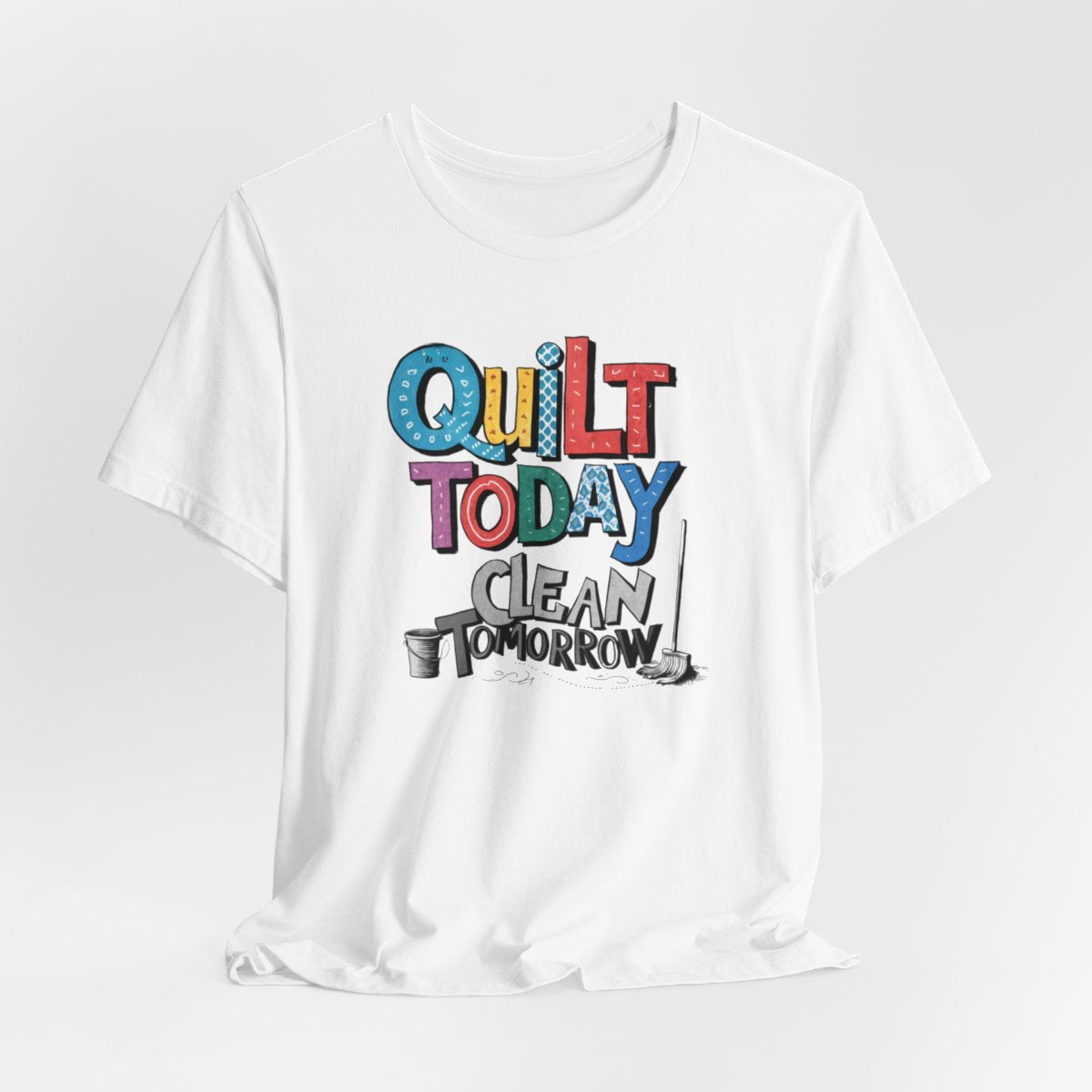 A White funny quilting T-Shirt with the phrase Quilt Today, Clean Tomorrow in a bold and cheerful font with a broom off to the side