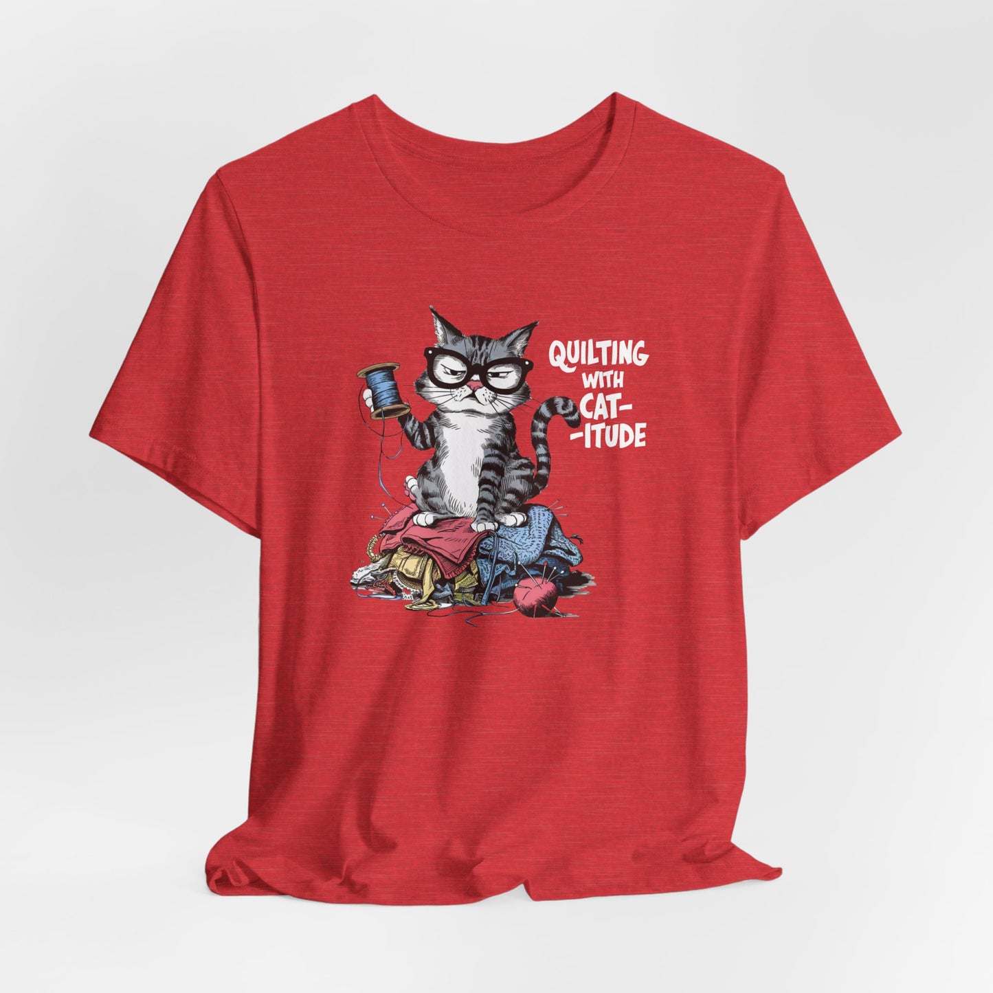 A Heather Red t-shirt featuring a sassy cartoon cat sitting on a pile of colorful fabric, holding a spool of thread, with the phrase 'Quilting with Cat-itude,' perfect for quilters and cat lovers.