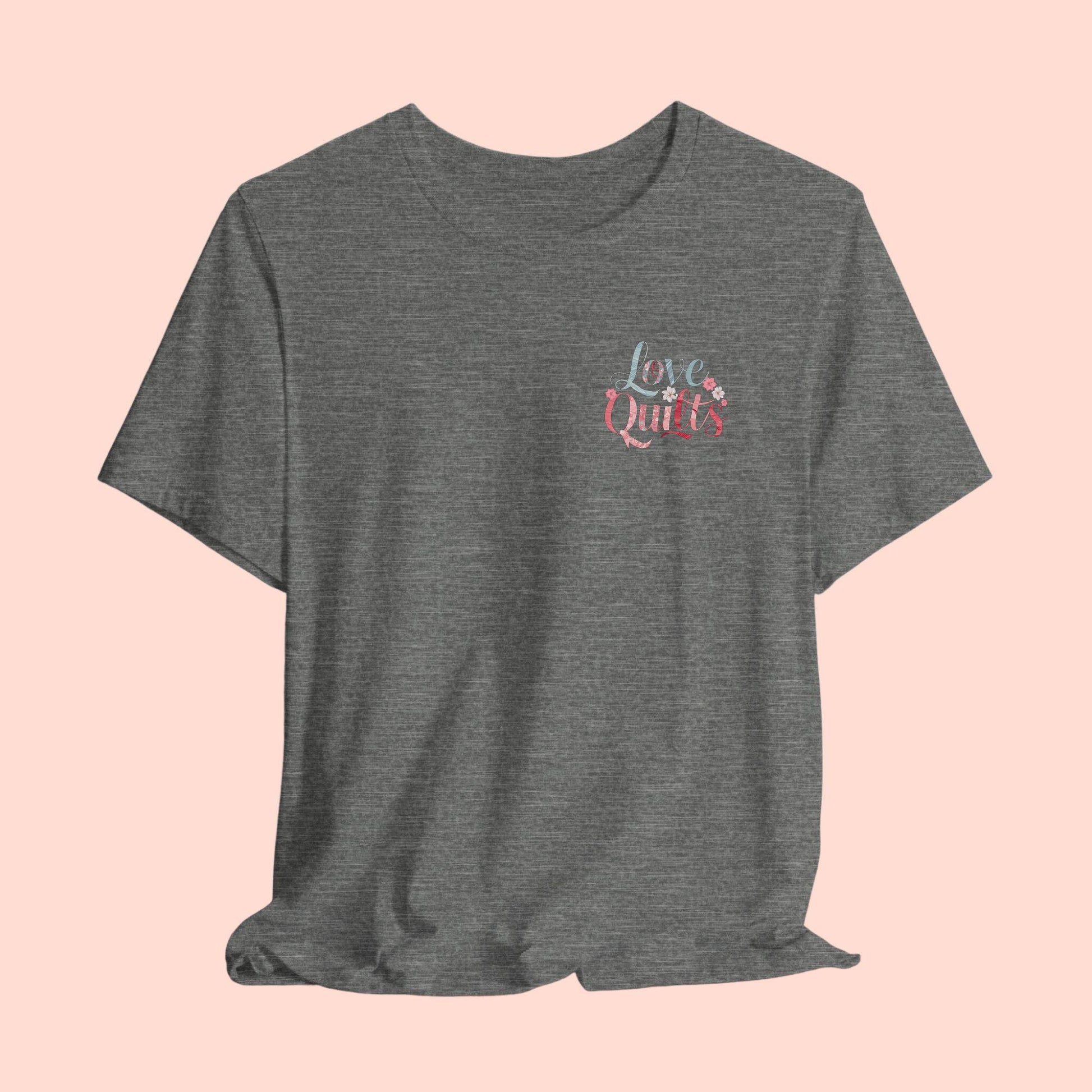 A t-shirt spelling out "Love Quilts". The word "Love" is in teal, while "Quilts" features various shades of pink and red. Small flowers in white and pink adorn the letters, creating a floral and quilting-inspired artwork.