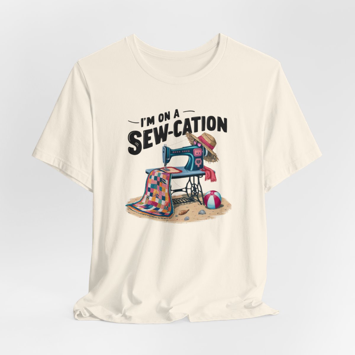 A Natural fun Quilting T-shirt of a vintage sewing machine on a beach with a colorful quilt, sun hat, and beach ball. Text above reads "I'm on a Sew-cation" in bold letters.