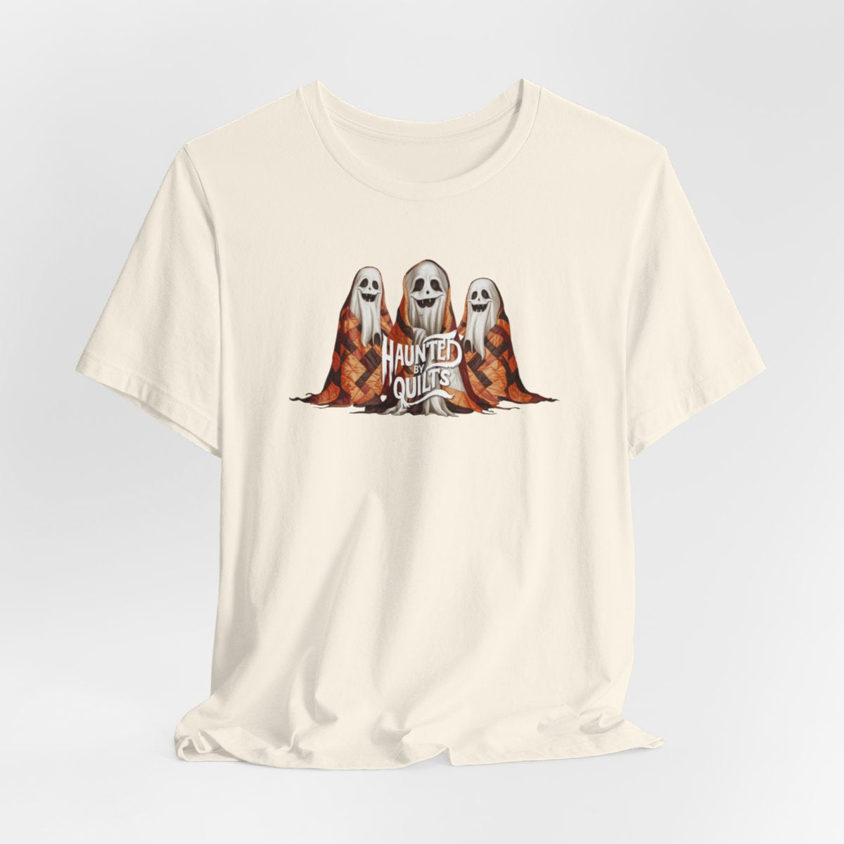 A Natural T-Shirt of three smiling cartoon ghosts covered in colorful patchwork quilts. The quilts feature orange, brown, and black patterns. Text below reads "HAUNTED BY QUILTS" in a spooky white font.