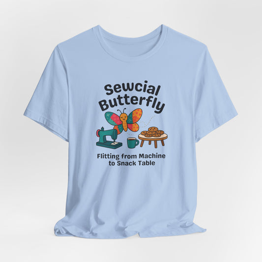 A A Baby Blue t-shirt with a colorful design showing a patchwork butterfly flying between a sewing machine and a table of cookies. Text reads “Sewcial Butterfly – Flitting from Machine to Snack Table.” Funny gift for quilters