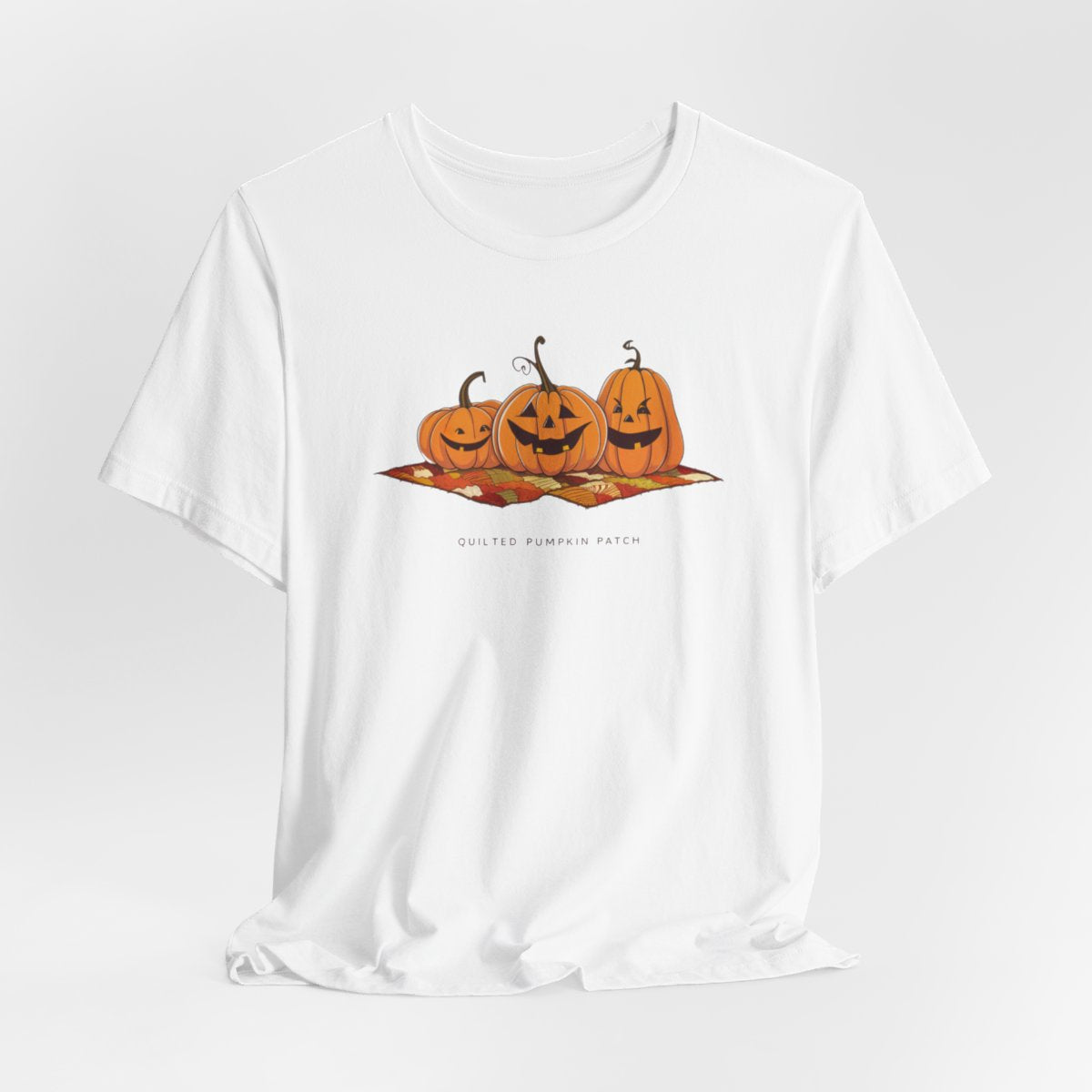 A White T-shirt with three smiling jack-o'-lanterns sitting on a colorful autumn-themed quilt. The pumpkins have carved faces with various expressions, and the quilt beneath them features a patchwork of fall colors. Text below reads "QUILTED PUMPKIN PATCH".