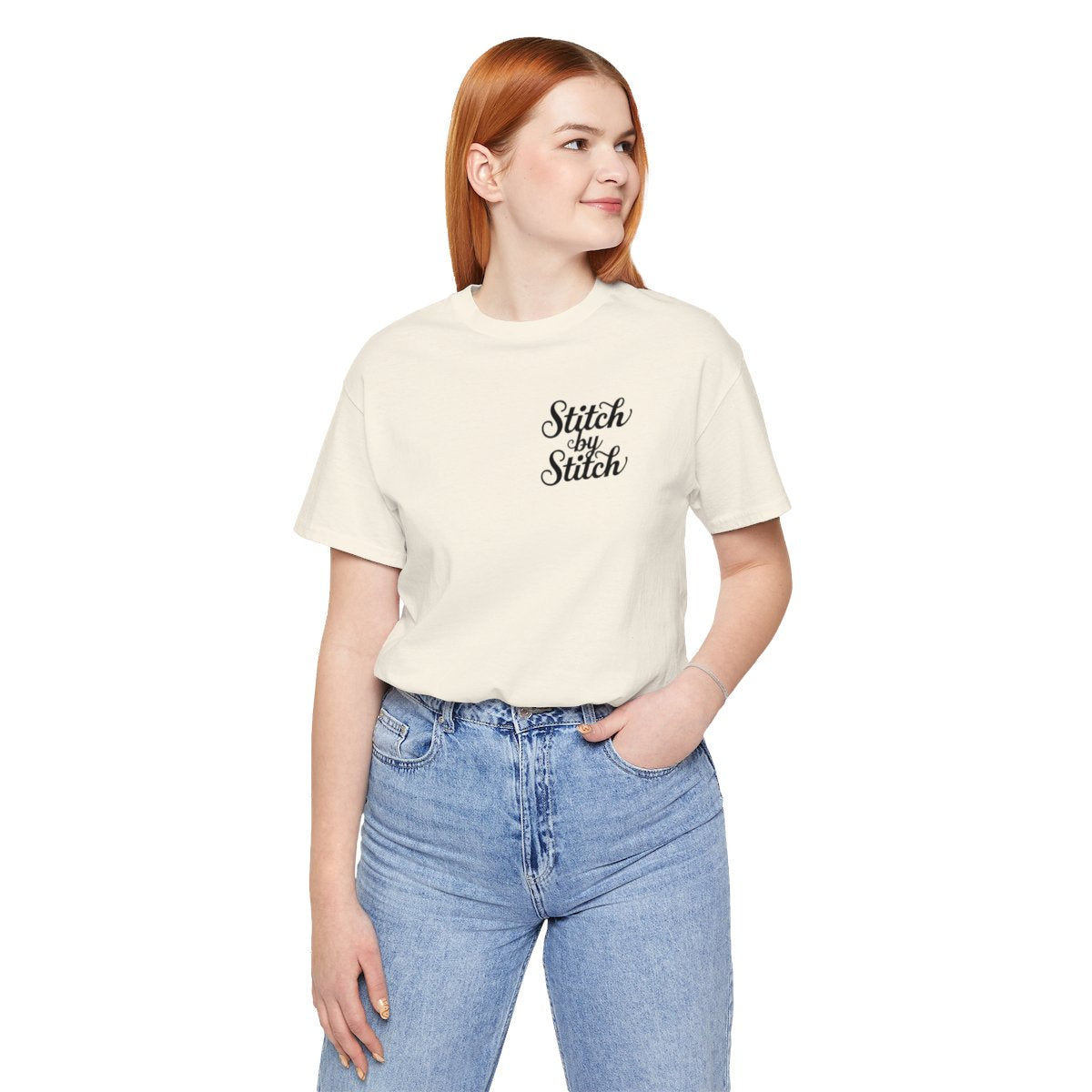 A Natural T-Shirt featuring the phrase "Stitch by Stitch". The word "Stitch" is written twice in a flowing script font, while "by" is centered between them in a smaller, sans-serif font. The design has a clean, minimalist aesthetic.