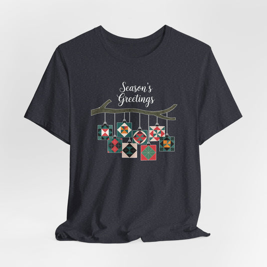 A Heather Navy christmas quilting t-shirt featuring quilt block ornaments hanging from a branch, featuring the phrase "Season's Greetings" above. The ornaments showcase various quilting patterns to highlight the holiday quilting theme.