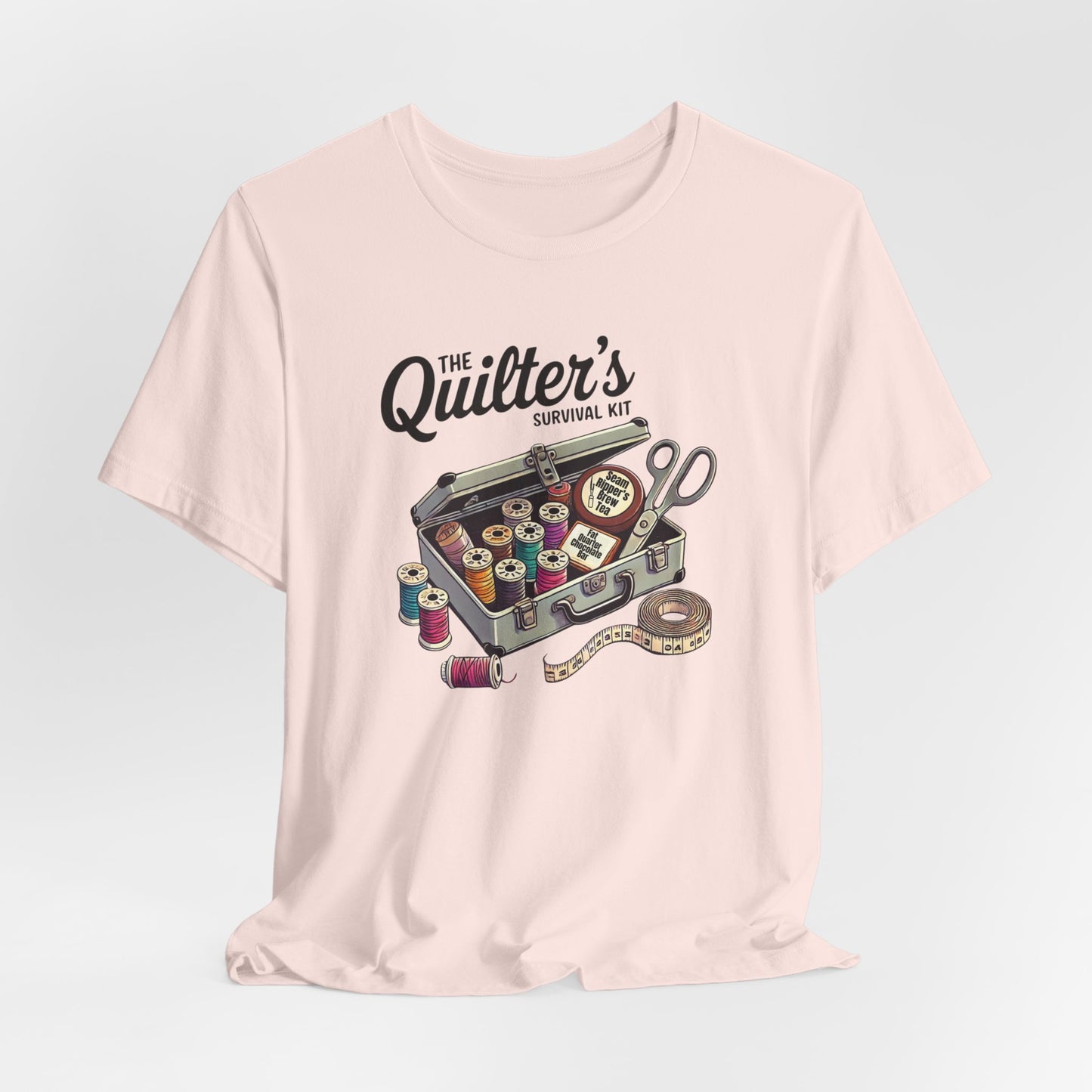 A Soft Pink t-shirt featuring a whimsical illustration of a quilter’s survival kit with colorful thread spools, scissors, and a tape measure, perfect for sewing and quilting enthusiasts.