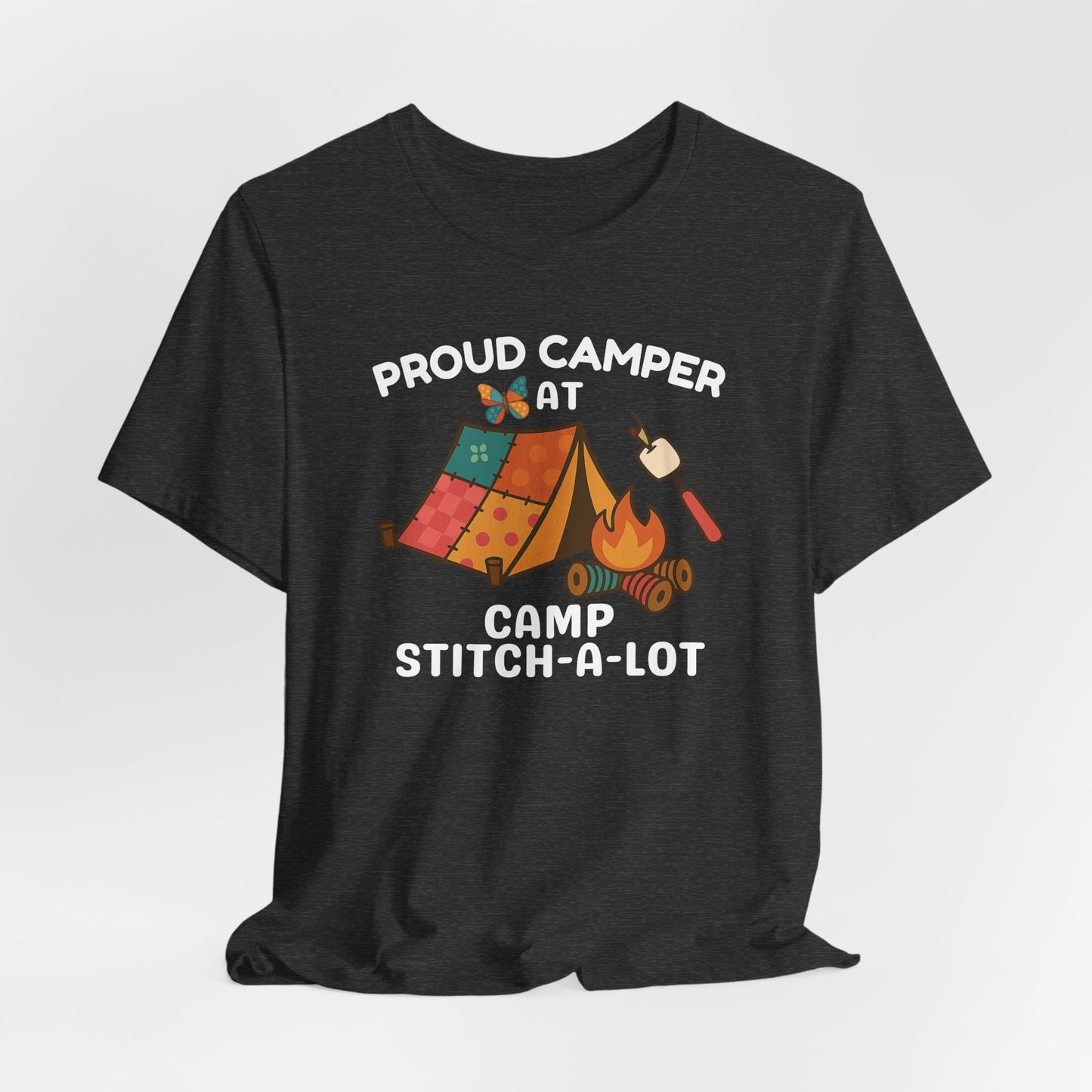 A A Dark Grey Heather t-shirt featuring a colorful design with a patchwork tent, thread spool campfire, marshmallow on a seam ripper stick, and a butterfly. Text reads "Proud Camper at Camp Stitch-a-Lot." Fun gift for quilting retreats.
