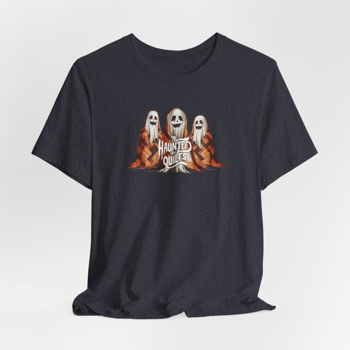 A Heather Navy T-Shirt of three smiling cartoon ghosts covered in colorful patchwork quilts. The quilts feature orange, brown, and black patterns. Text below reads "HAUNTED BY QUILTS" in a spooky white font.