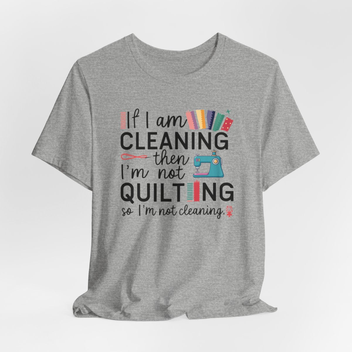 An Athletic Heather funny quilting T-Shirt with the phrase If I am Cleaning then I'm not Quilting in a bold and cheerful font with images of quilting icons and supplies