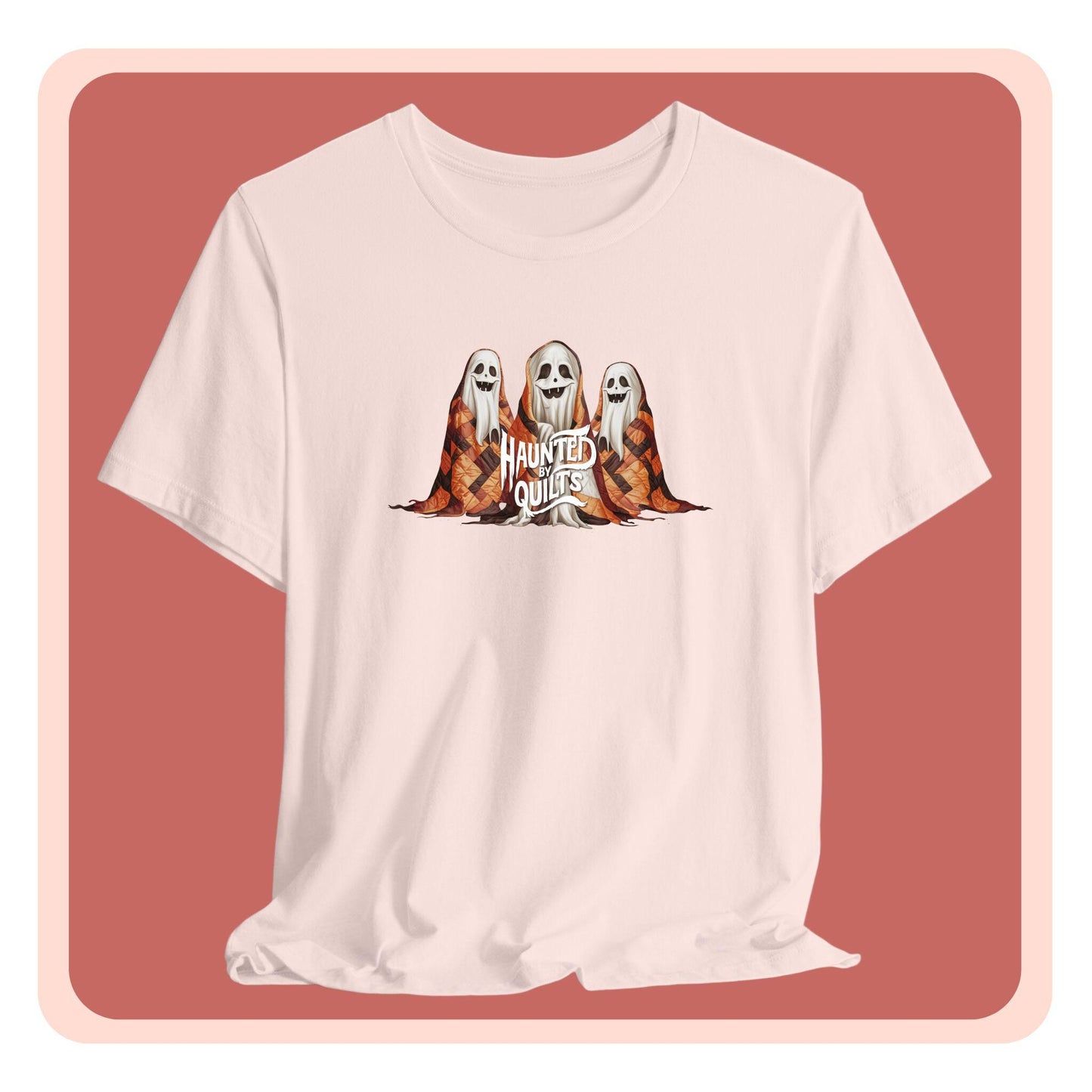 A T-Shirt of three smiling cartoon ghosts covered in colorful patchwork quilts. The quilts feature orange, brown, and black patterns. Text below reads "HAUNTED BY QUILTS" in a spooky white font.