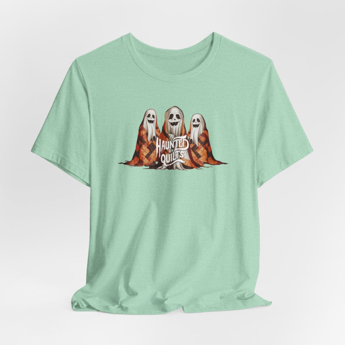 A Heather Mint T-Shirt of three smiling cartoon ghosts covered in colorful patchwork quilts. The quilts feature orange, brown, and black patterns. Text below reads "HAUNTED BY QUILTS" in a spooky white font.