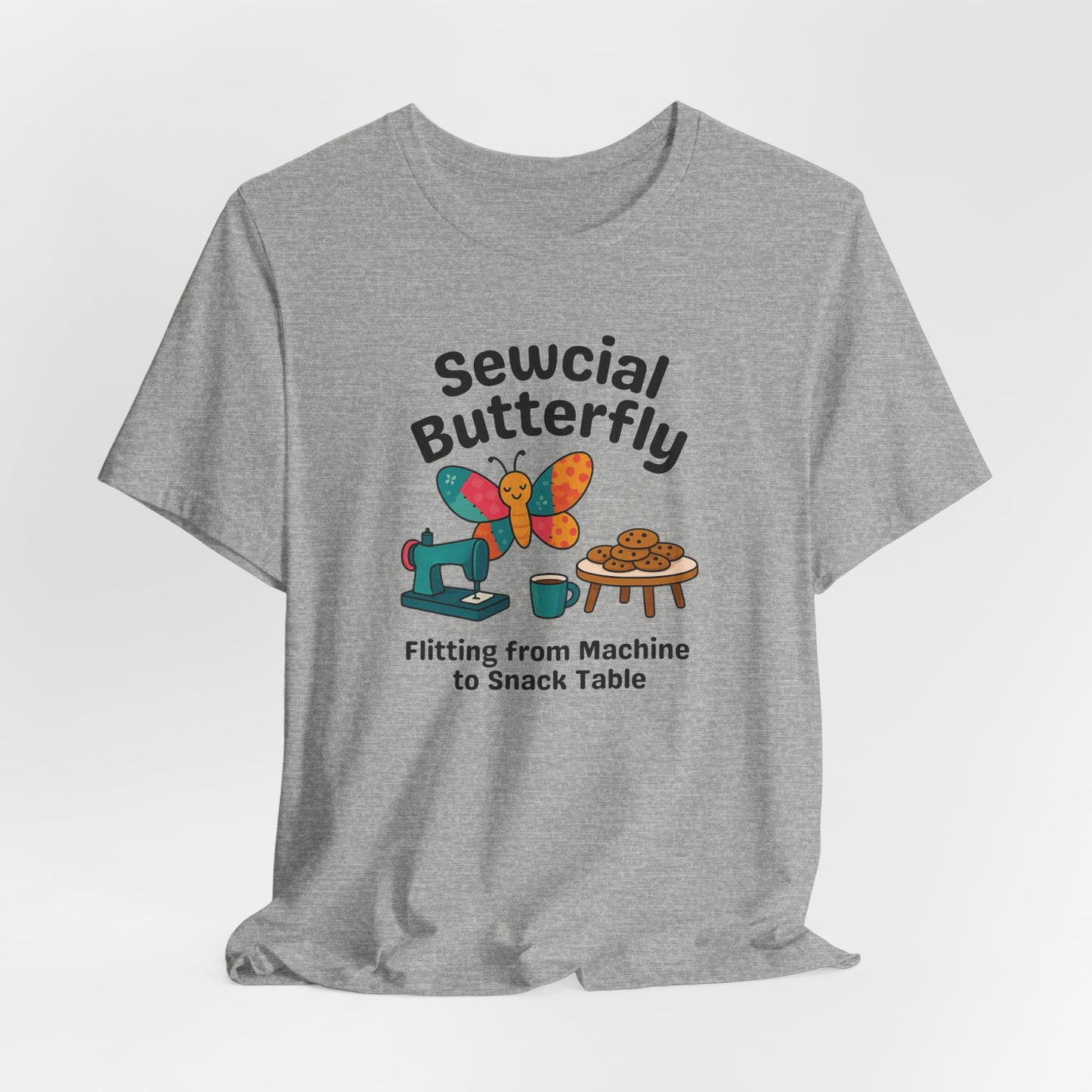 A An Athletic Heather t-shirt with a colorful design showing a patchwork butterfly flying between a sewing machine and a table of cookies. Text reads “Sewcial Butterfly – Flitting from Machine to Snack Table.” Funny gift for quilters