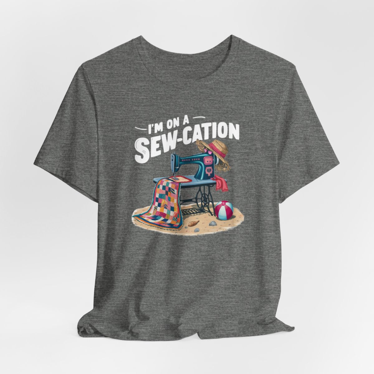 A Deep Heather fun Quilting T-shirt of a vintage sewing machine on a beach with a colorful quilt, sun hat, and beach ball. Text above reads "I'm on a Sew-cation" in bold letters.