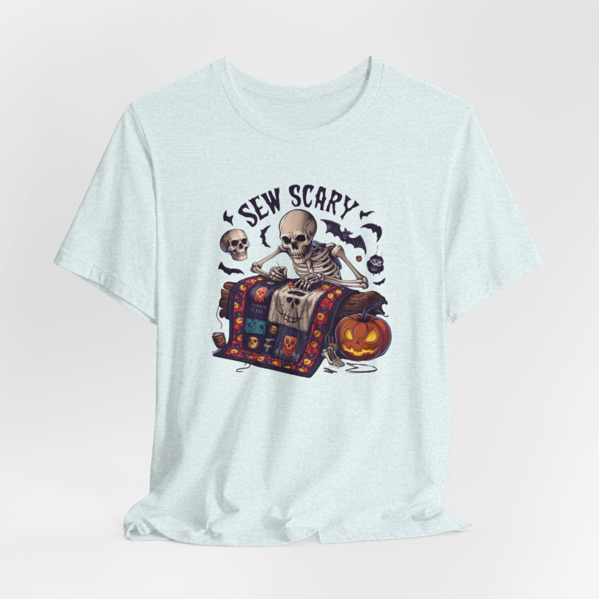 A Heather Ice Blue Halloween Quilting T-Shirt featuring a skeleton sewing Halloween quilt. Colorful patchwork with spooky motifs. Bats and pumpkins surround skeleton. Text reads "Sew Scary". Ideal for quilters who love Halloween.