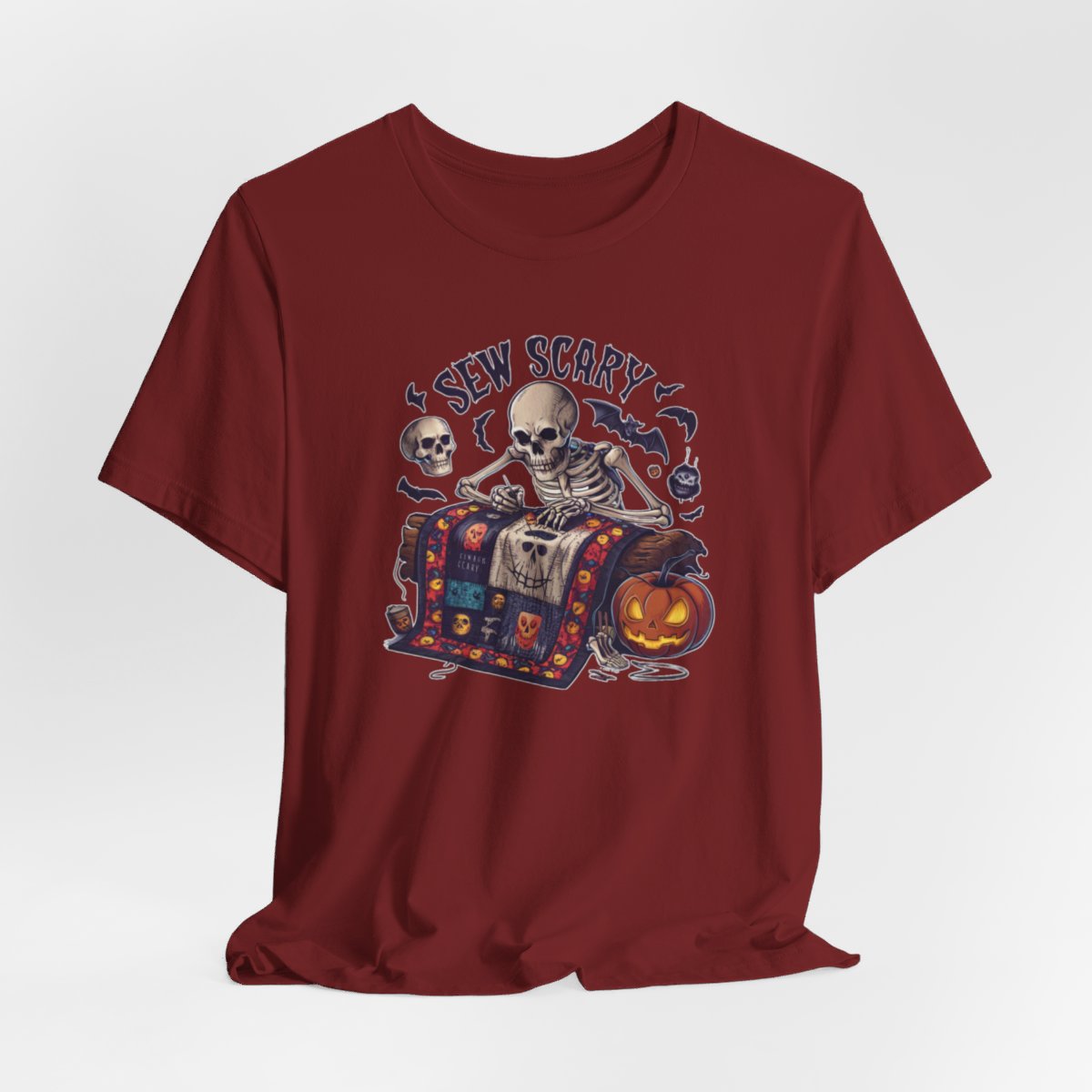 A Cardinal Halloween Quilting T-Shirt featuring a skeleton sewing Halloween quilt. Colorful patchwork with spooky motifs. Bats and pumpkins surround skeleton. Text reads "Sew Scary". Ideal for quilters who love Halloween.
