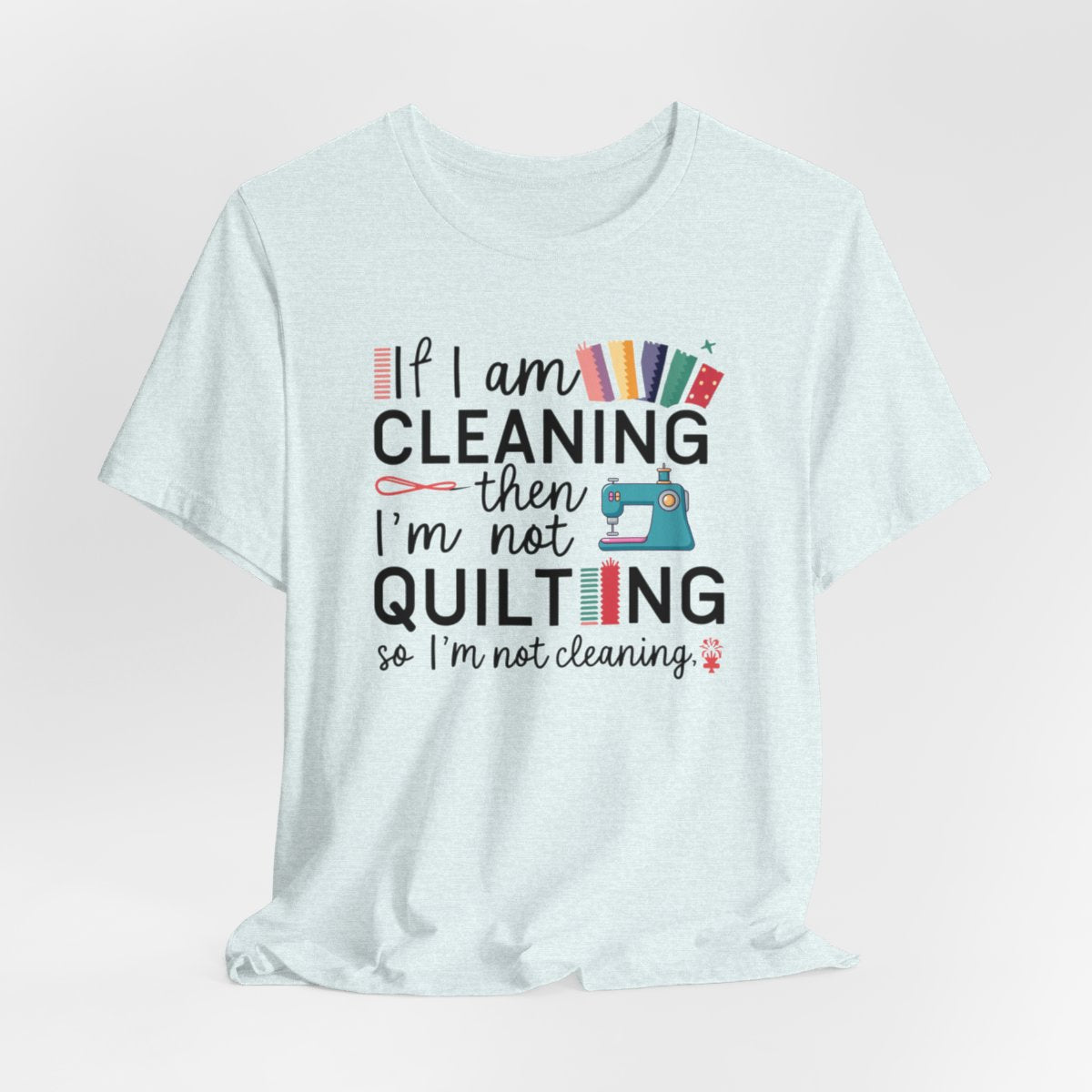 A Heather Ice Blue funny quilting T-Shirt with the phrase If I am Cleaning then I'm not Quilting in a bold and cheerful font with images of quilting icons and supplies