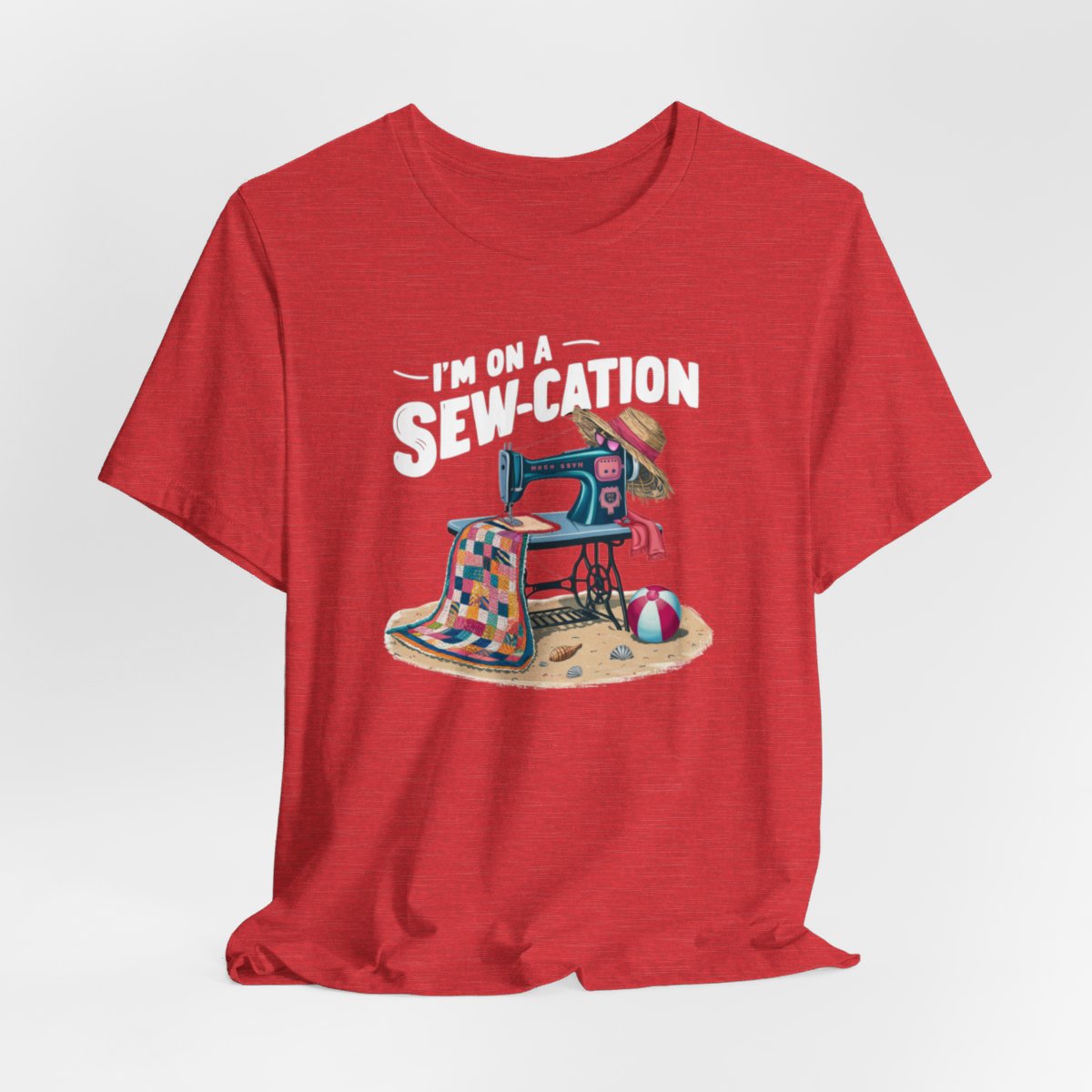 A Heather Red fun Quilting T-shirt of a vintage sewing machine on a beach with a colorful quilt, sun hat, and beach ball. Text above reads "I'm on a Sew-cation" in bold letters.