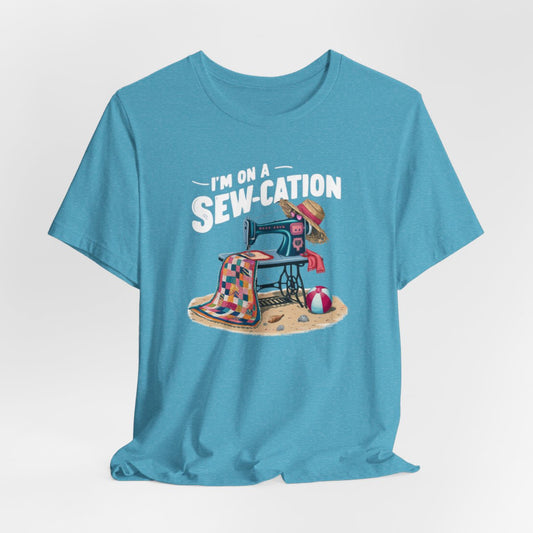 A Heather Aqua fun Quilting T-shirt of a vintage sewing machine on a beach with a colorful quilt, sun hat, and beach ball. Text above reads "I'm on a Sew-cation" in bold letters.