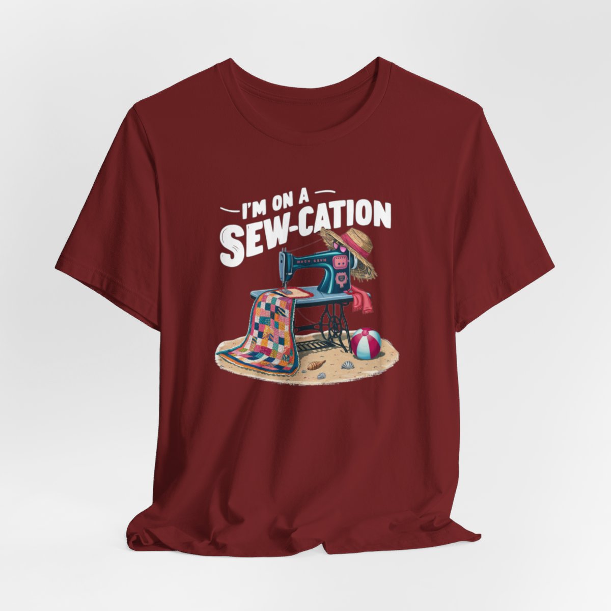 A Cardinal fun Quilting T-shirt of a vintage sewing machine on a beach with a colorful quilt, sun hat, and beach ball. Text above reads "I'm on a Sew-cation" in bold letters.