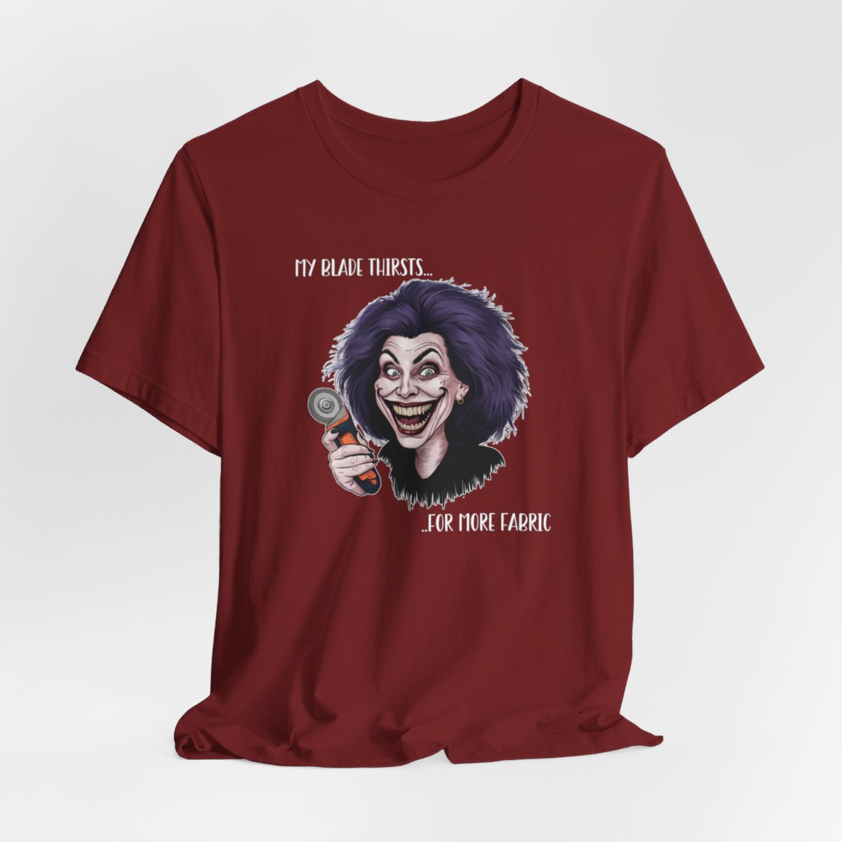 A Cardinal T-shirt showing a cartoonish character with wild purple hair and an exaggerated grin, holding a rotary cutter. Text reads "My blade thirsts... for more fabric", humorously depicting a quilter's endless desire for new materials.