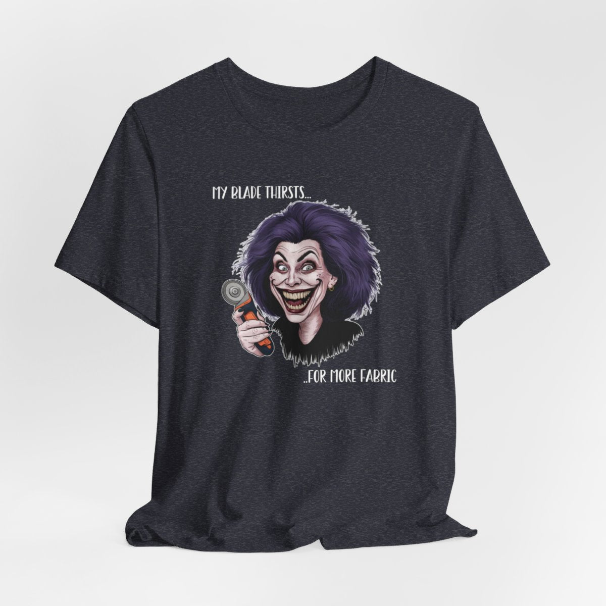 A Heather Navy T-shirt showing a cartoonish character with wild purple hair and an exaggerated grin, holding a rotary cutter. Text reads "My blade thirsts... for more fabric", humorously depicting a quilter's endless desire for new materials.
