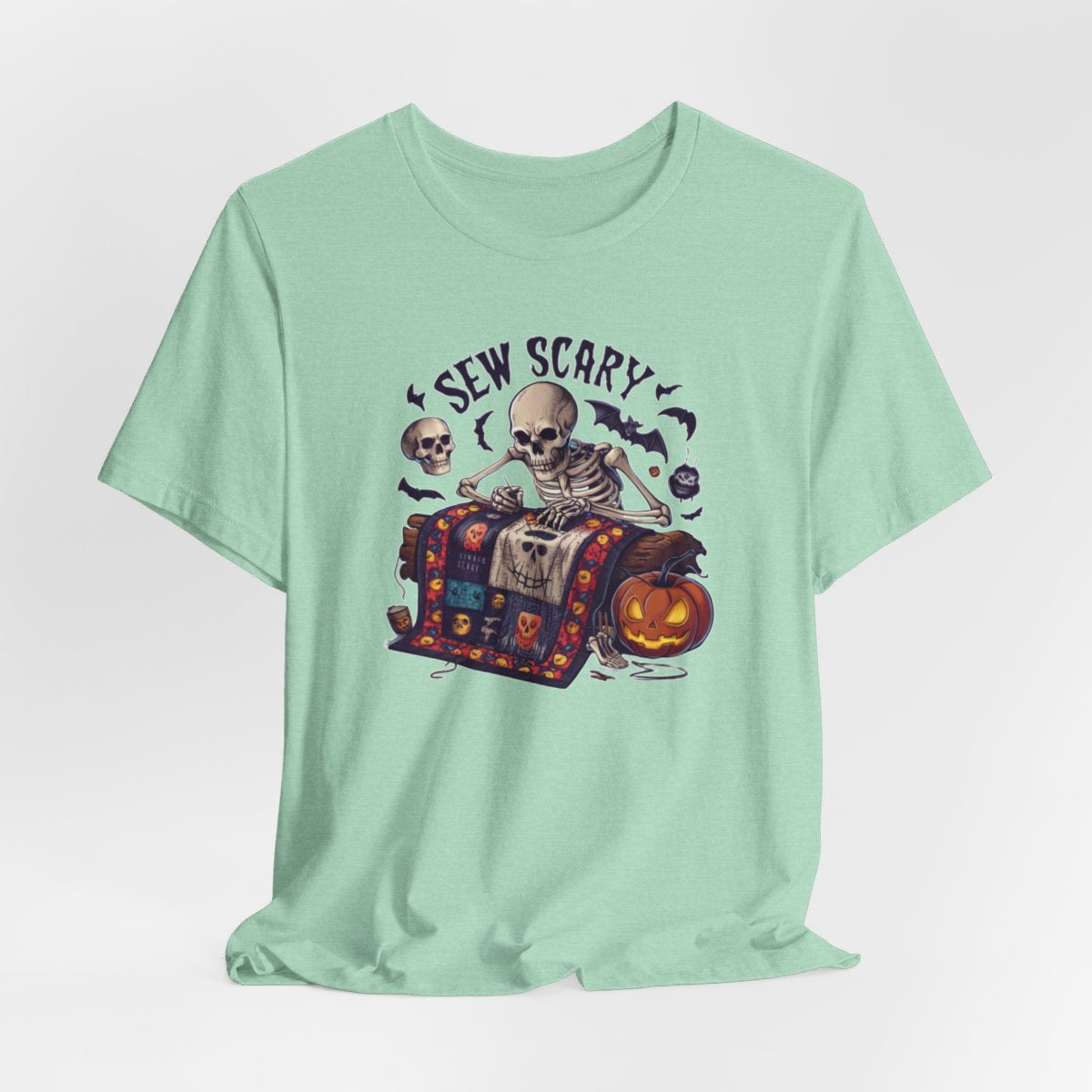 A Heather Mint Halloween Quilting T-Shirt featuring a skeleton sewing Halloween quilt. Colorful patchwork with spooky motifs. Bats and pumpkins surround skeleton. Text reads "Sew Scary". Ideal for quilters who love Halloween.