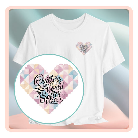 A T-shirt showing a large heart-shaped quilt pattern in pastel colors of pink, blue, and cream. Black text overlaid on the heart reads "Quilters make the world a softer place" in a mix of script and sans-serif fonts. The design combines geometric quilt patterns with an uplifting message about quilting.