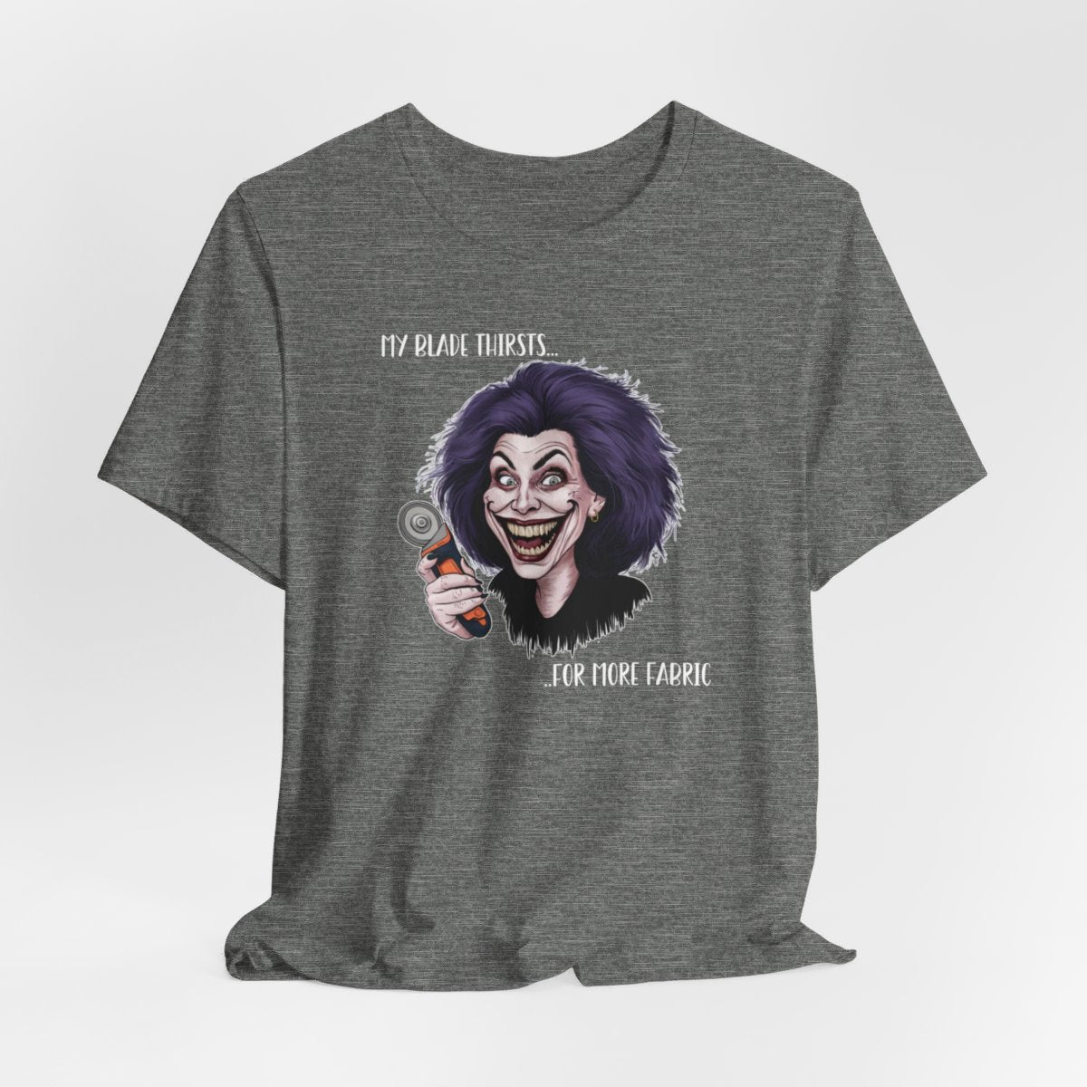 A Deep Heather T-shirt showing a cartoonish character with wild purple hair and an exaggerated grin, holding a rotary cutter. Text reads "My blade thirsts... for more fabric", humorously depicting a quilter's endless desire for new materials.