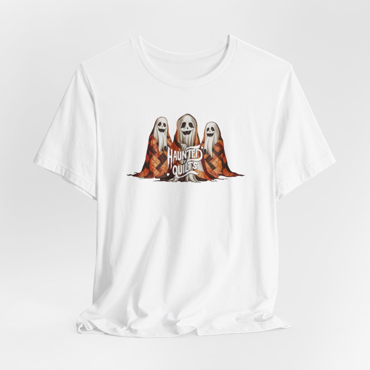 A White T-Shirt of three smiling cartoon ghosts covered in colorful patchwork quilts. The quilts feature orange, brown, and black patterns. Text below reads "HAUNTED BY QUILTS" in a spooky white font.