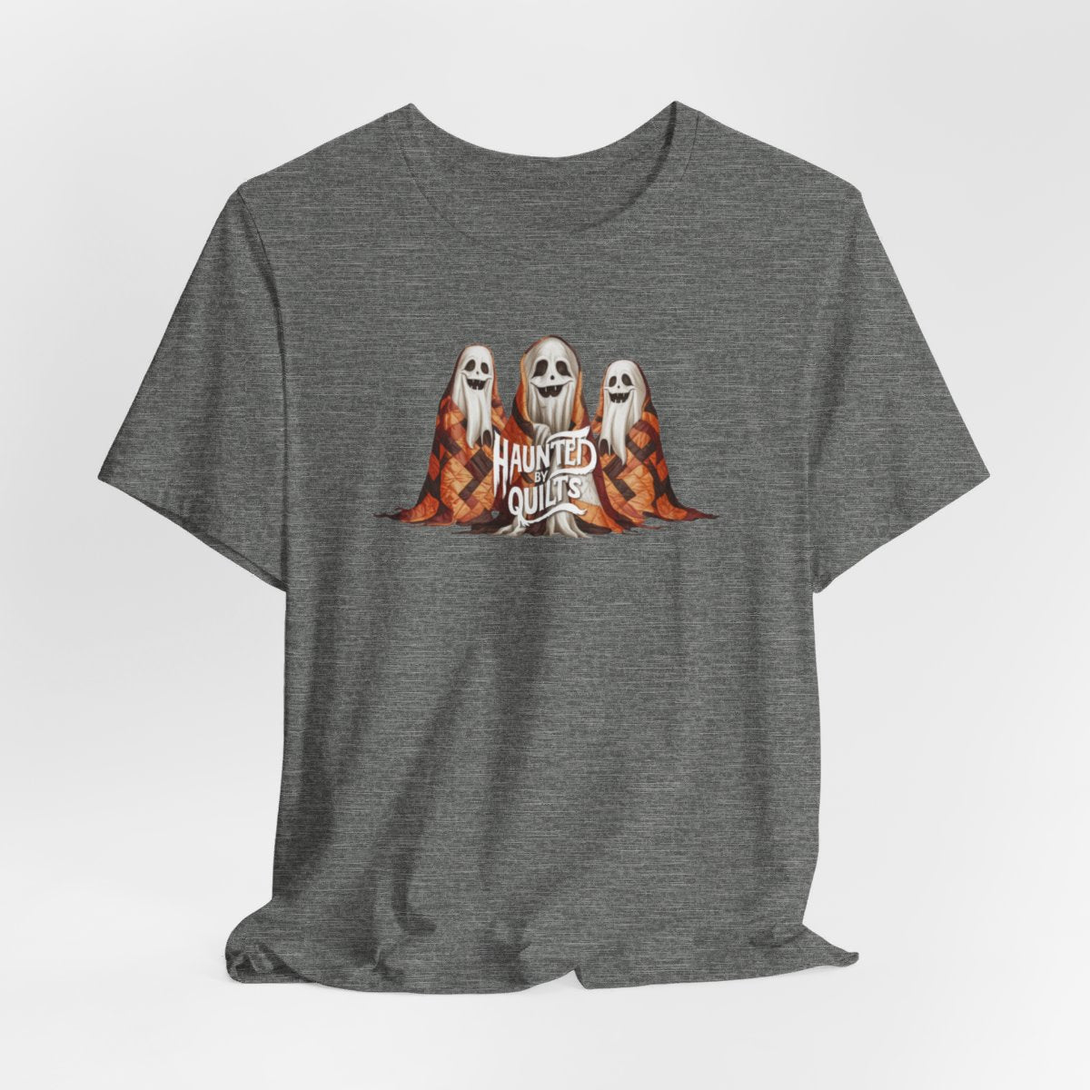 A Deep Heather T-Shirt of three smiling cartoon ghosts covered in colorful patchwork quilts. The quilts feature orange, brown, and black patterns. Text below reads "HAUNTED BY QUILTS" in a spooky white font.