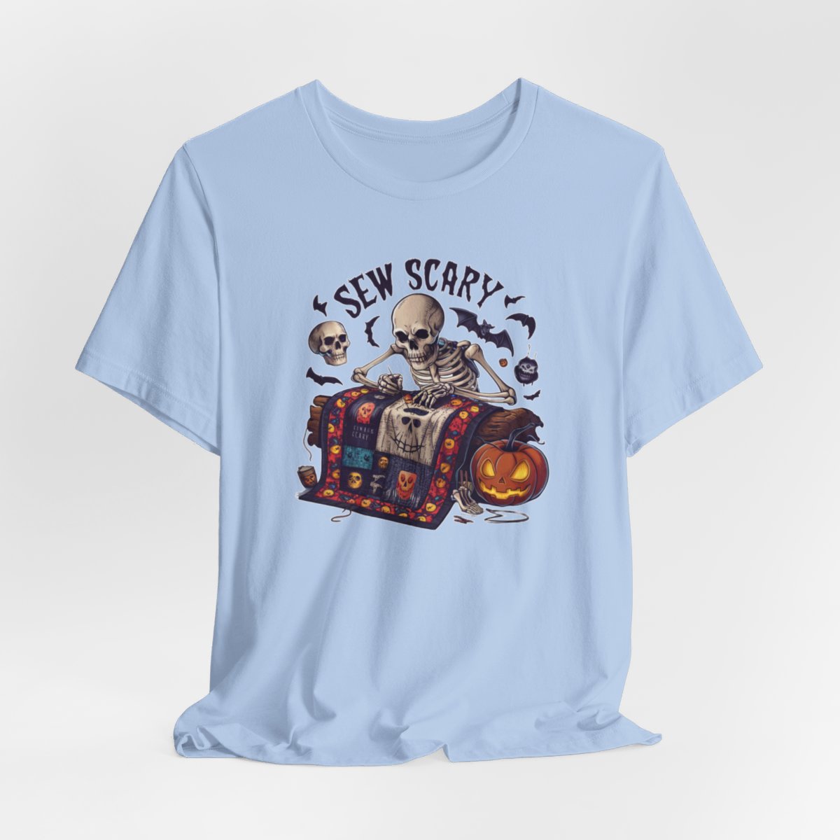 A Baby Blue Halloween Quilting T-Shirt featuring a skeleton sewing Halloween quilt. Colorful patchwork with spooky motifs. Bats and pumpkins surround skeleton. Text reads "Sew Scary". Ideal for quilters who love Halloween.