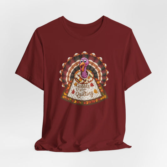 A Cardinal thanksgiving  quilting t-shirt featuring a quilted turkey design with colorful patchwork feathers and body. Text on turkey reads "Gobble up some Quilting" surrounded by fall leaves. Combines Thanksgiving imagery with quilting motifs in a whimsical illustration.