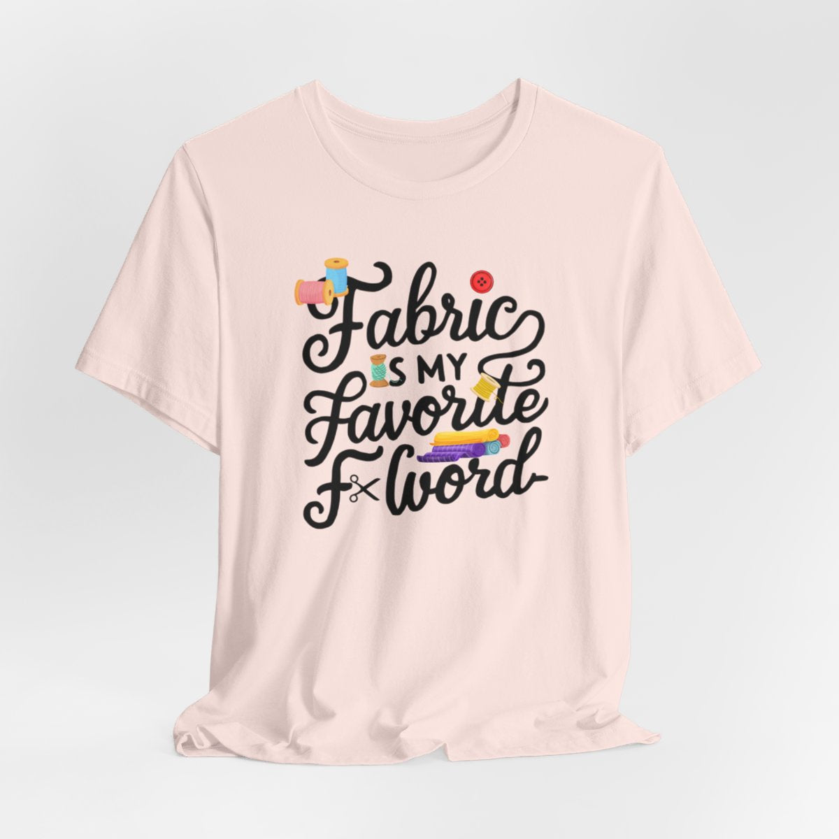 A Soft Pink T-Shirt with the phrase Fabric is My Favorite F-Word in a bold font with thread, buttons, and fabric bolts