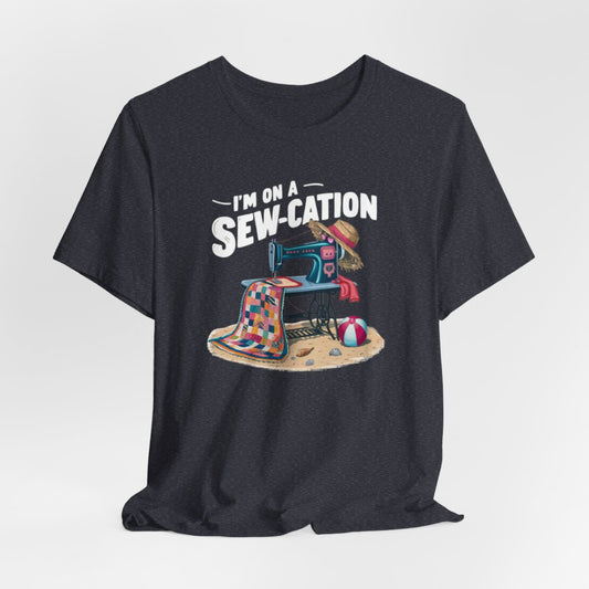 A Heather Navy fun Quilting T-shirt of a vintage sewing machine on a beach with a colorful quilt, sun hat, and beach ball. Text above reads "I'm on a Sew-cation" in bold letters.