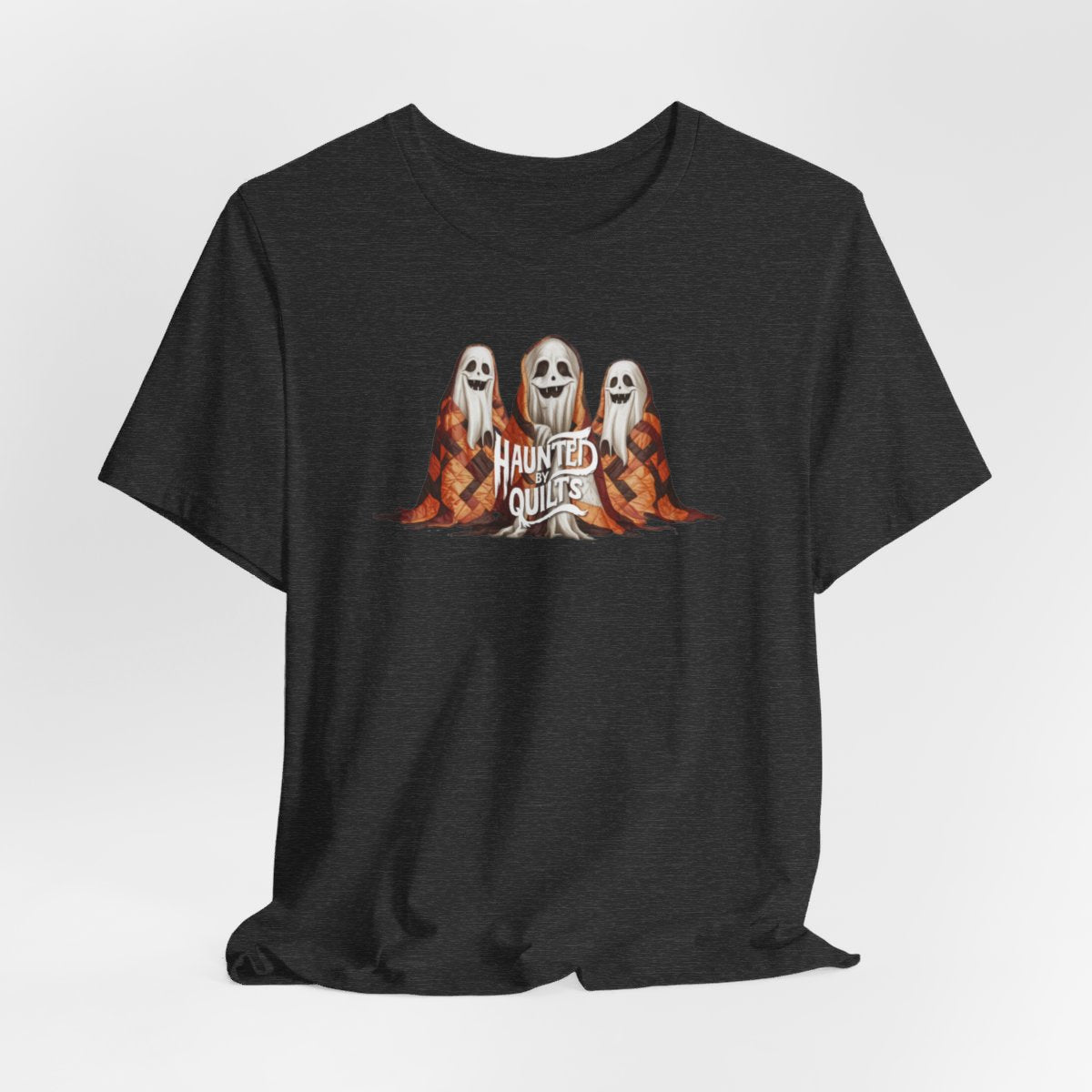 A Dark Grey Heather T-Shirt of three smiling cartoon ghosts covered in colorful patchwork quilts. The quilts feature orange, brown, and black patterns. Text below reads "HAUNTED BY QUILTS" in a spooky white font.
