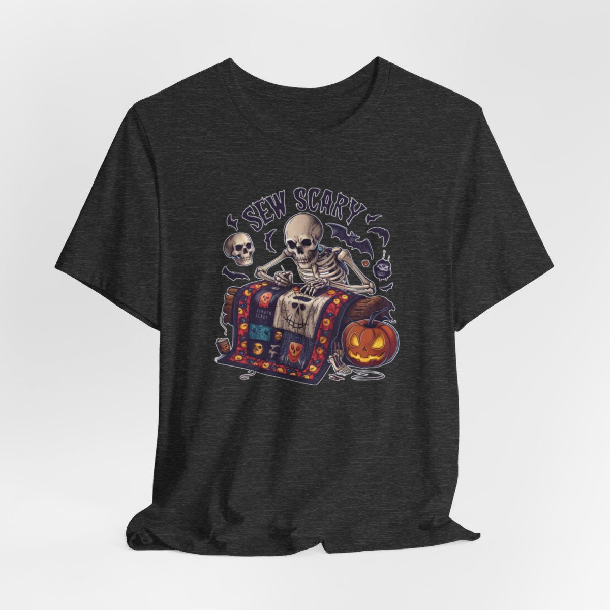 A Dark Grey Heather Halloween Quilting T-Shirt featuring a skeleton sewing Halloween quilt. Colorful patchwork with spooky motifs. Bats and pumpkins surround skeleton. Text reads "Sew Scary". Ideal for quilters who love Halloween.
