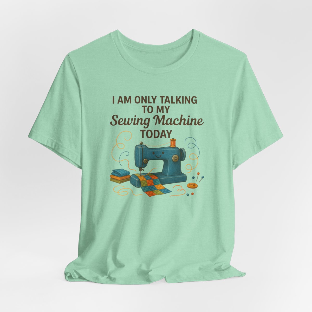 A Heather Mint Short Sleeve T-Shirt featuring a smiling sewing machine with colorful fabric and playful thread swirls