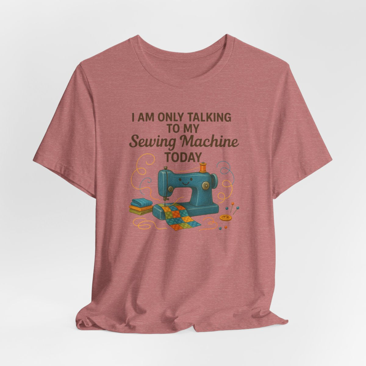 A Heather Mauve Short Sleeve T-Shirt featuring a smiling sewing machine with colorful fabric and playful thread swirls