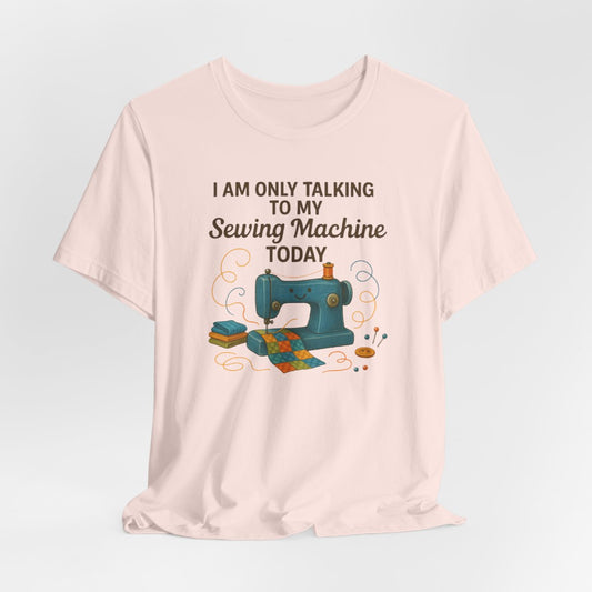 A Soft Pink Short Sleeve T-Shirt featuring a smiling sewing machine with colorful fabric and playful thread swirls