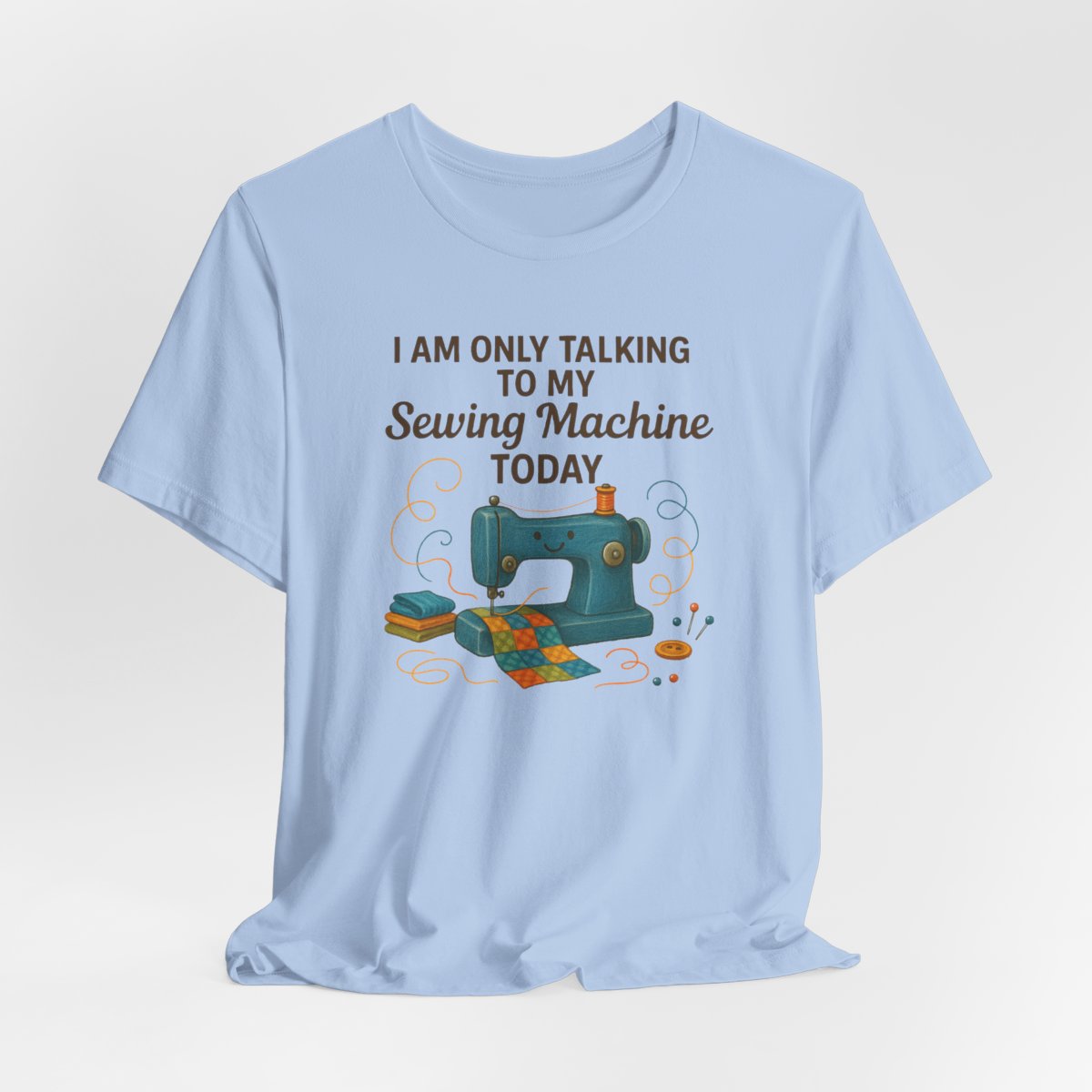 A Baby Blue Short Sleeve T-Shirt featuring a smiling sewing machine with colorful fabric and playful thread swirls