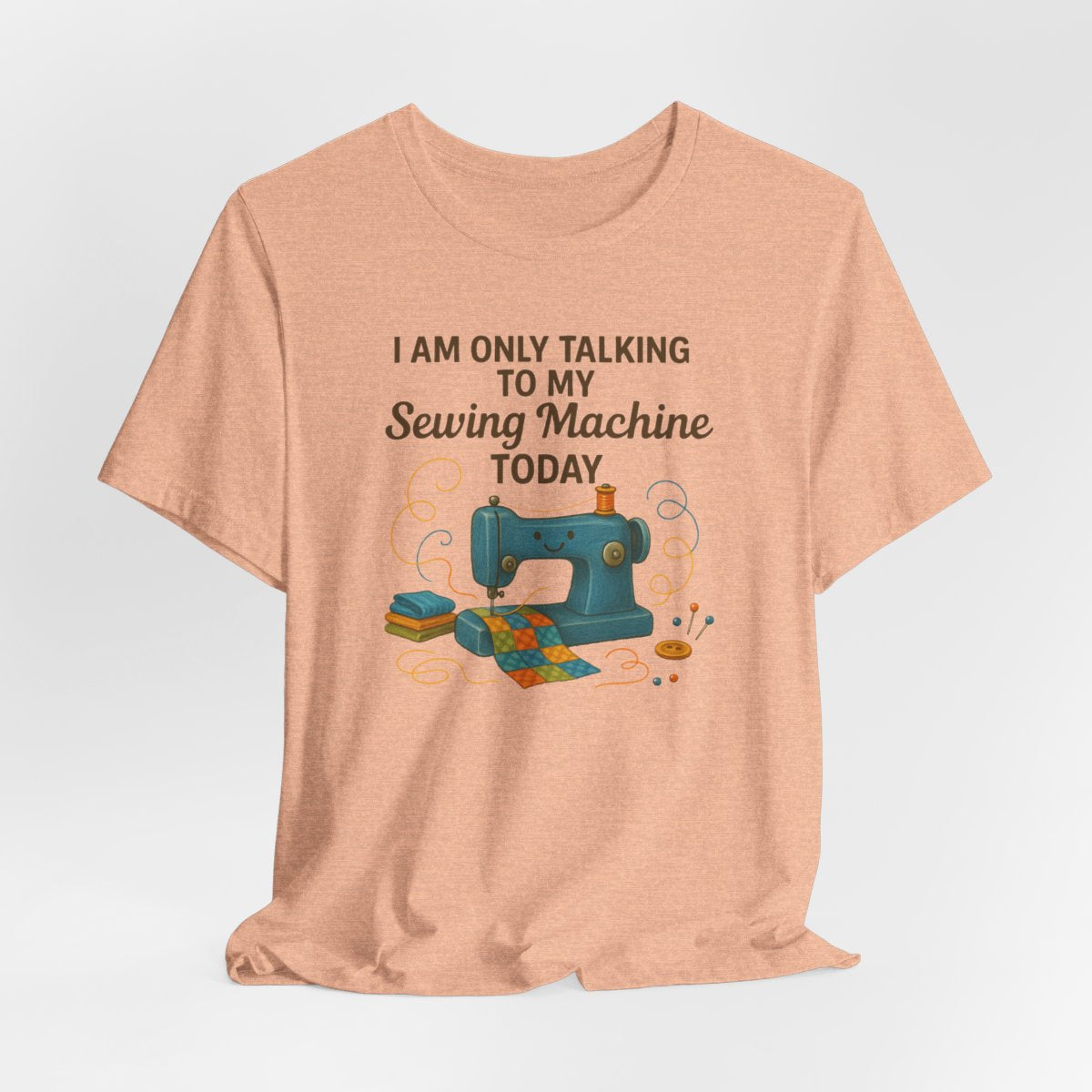 A Heather Peach Short Sleeve T-Shirt featuring a smiling sewing machine with colorful fabric and playful thread swirls