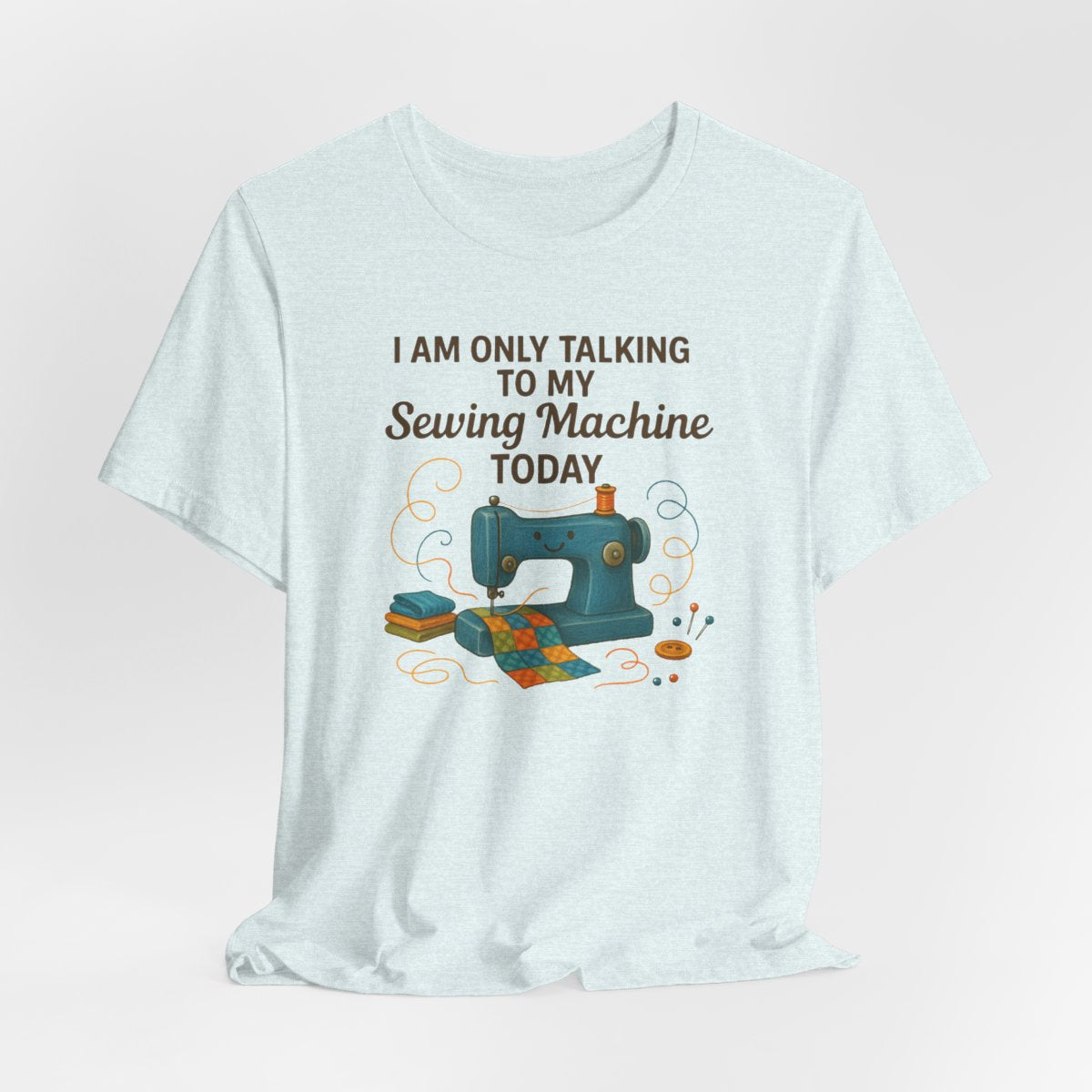 A Heather Ice Blue Short Sleeve T-Shirt featuring a smiling sewing machine with colorful fabric and playful thread swirls