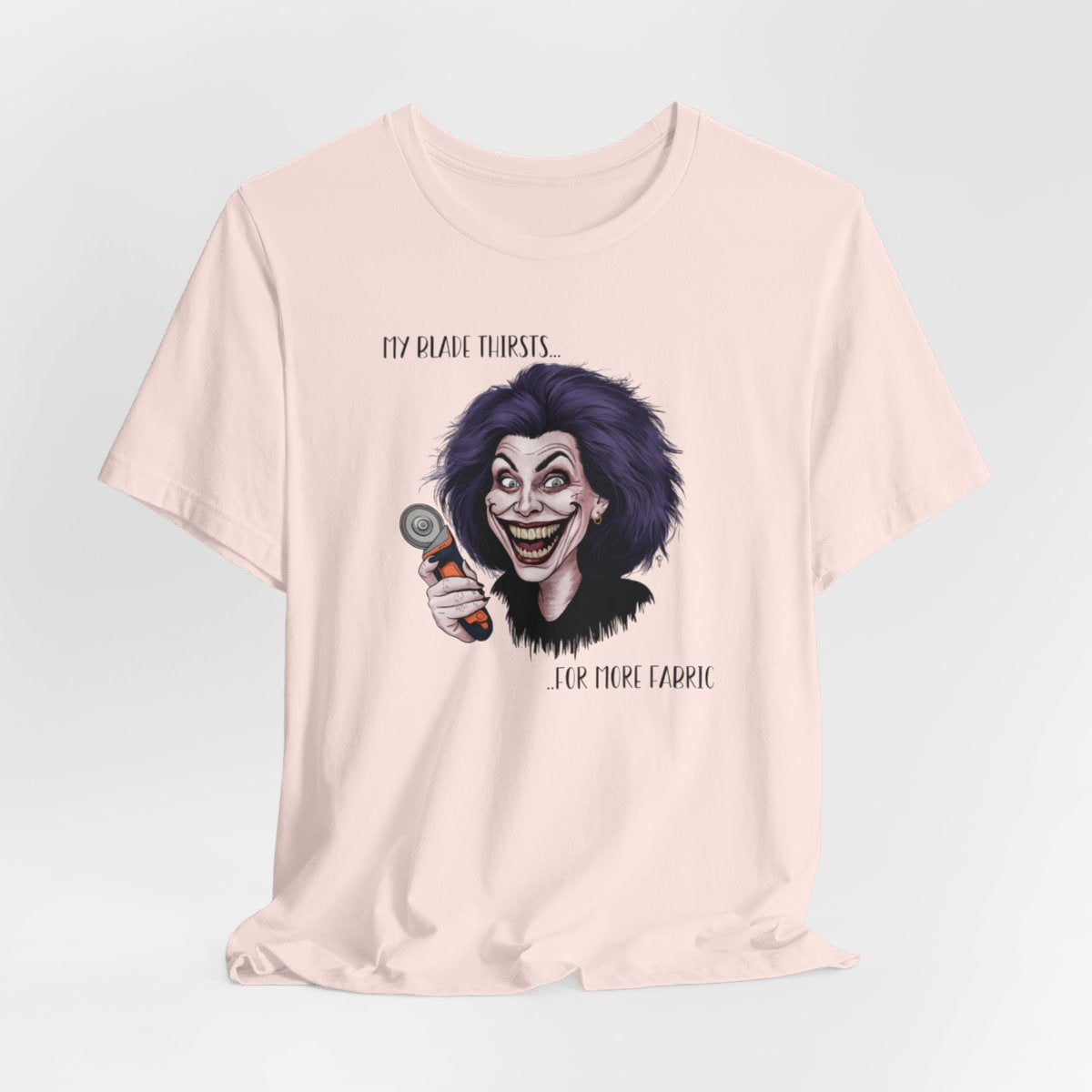 A Soft Pink T-shirt showing a cartoonish character with wild purple hair and an exaggerated grin, holding a rotary cutter. Text reads "My blade thirsts... for more fabric", humorously depicting a quilter's endless desire for new materials.